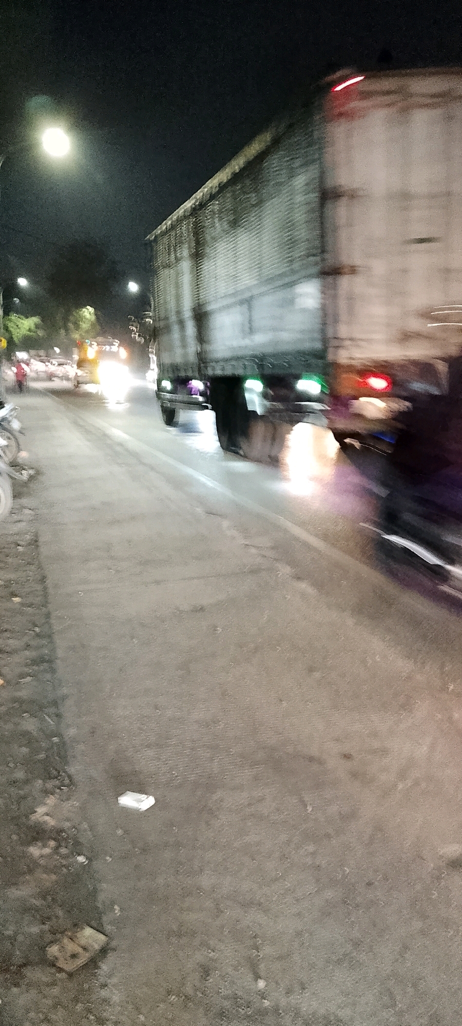 A blurry, nighttime photograph captures a street scene in Bekasi, Indonesia. A large truck is the primary subject, moving from left to right across the frame and creating motion blur. Its trailer appears to be a light color, possibly white or grey, with a dark-colored frame along the top. The truck has several lights visible, including green lights on the lower rear and a red light strip across the top. Flames or bright lights are also visible beneath the rear wheels of the truck, possibly from exhaust or road debris.
To the left of the truck, other vehicles are present, also exhibiting motion blur due to the nighttime setting and likely movement. One vehicle appears to be a lighter color with bright headlights, possibly a car or another truck. A motorcycle is partially visible in the foreground on the left. A few indistinct figures, possibly pedestrians, are also present in the distance on the left side of the road.
The environment appears to be an urban street. Lampposts are visible in the background, emitting light that illuminates the scene. The road surface is paved and appears to be somewhat worn or dirty. Debris, including what looks like a white rectangular object and some darker, flatter objects, are scattered on the road's shoulder. Trees are visible in the far background, silhouetted against the night sky. The overall impression is of a busy, active street at night, with a focus on the movement of the large truck.