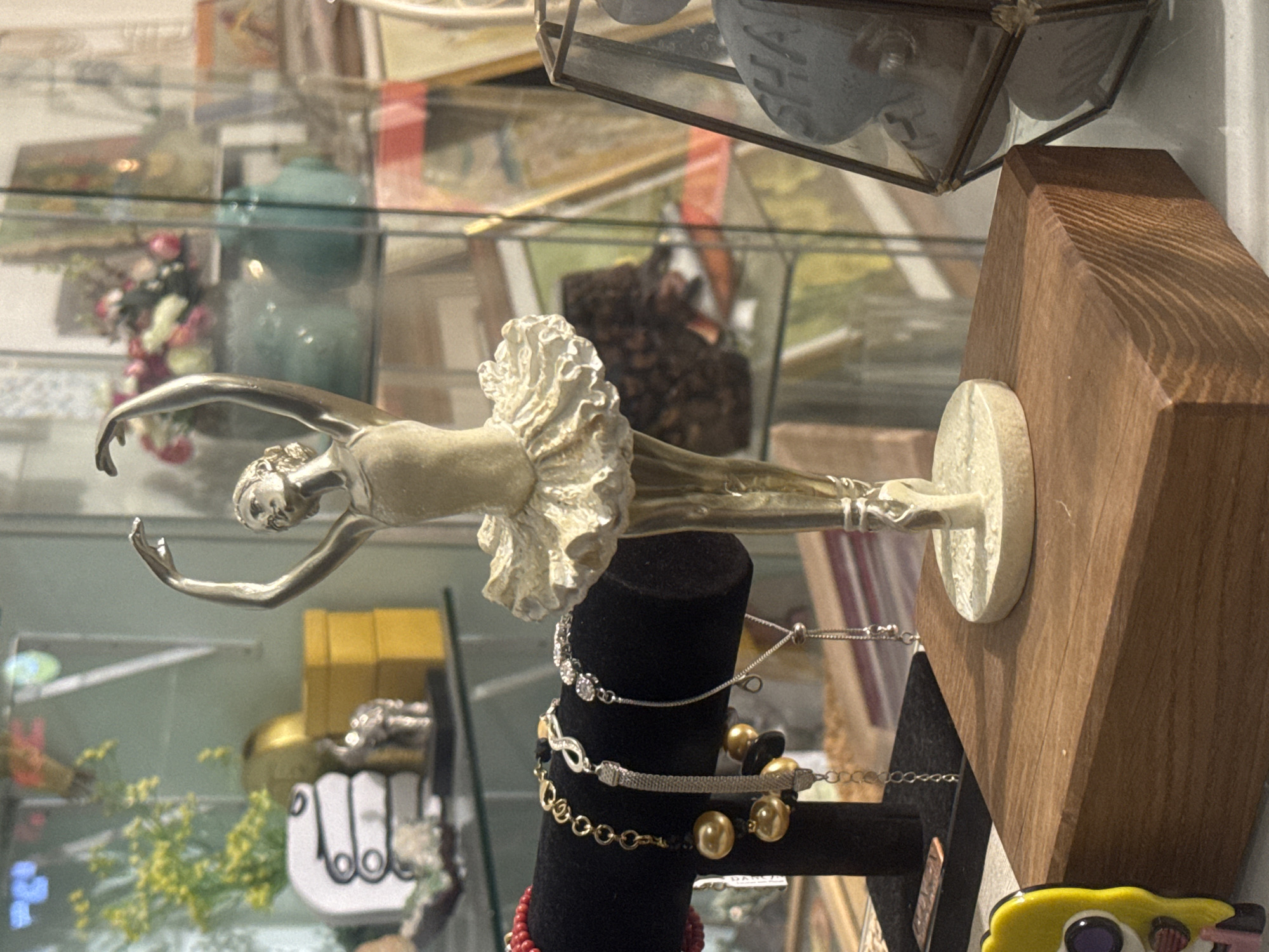 Observed in Givatayim, Israel, is a tableau featuring a silver-toned ballerina figurine with a white tutu positioned horizontally atop a black velvet jewelry display stand adorned with several bracelets. The jewelry stand itself rests against a wooden block. The scene is set within a glass display case containing various other items.