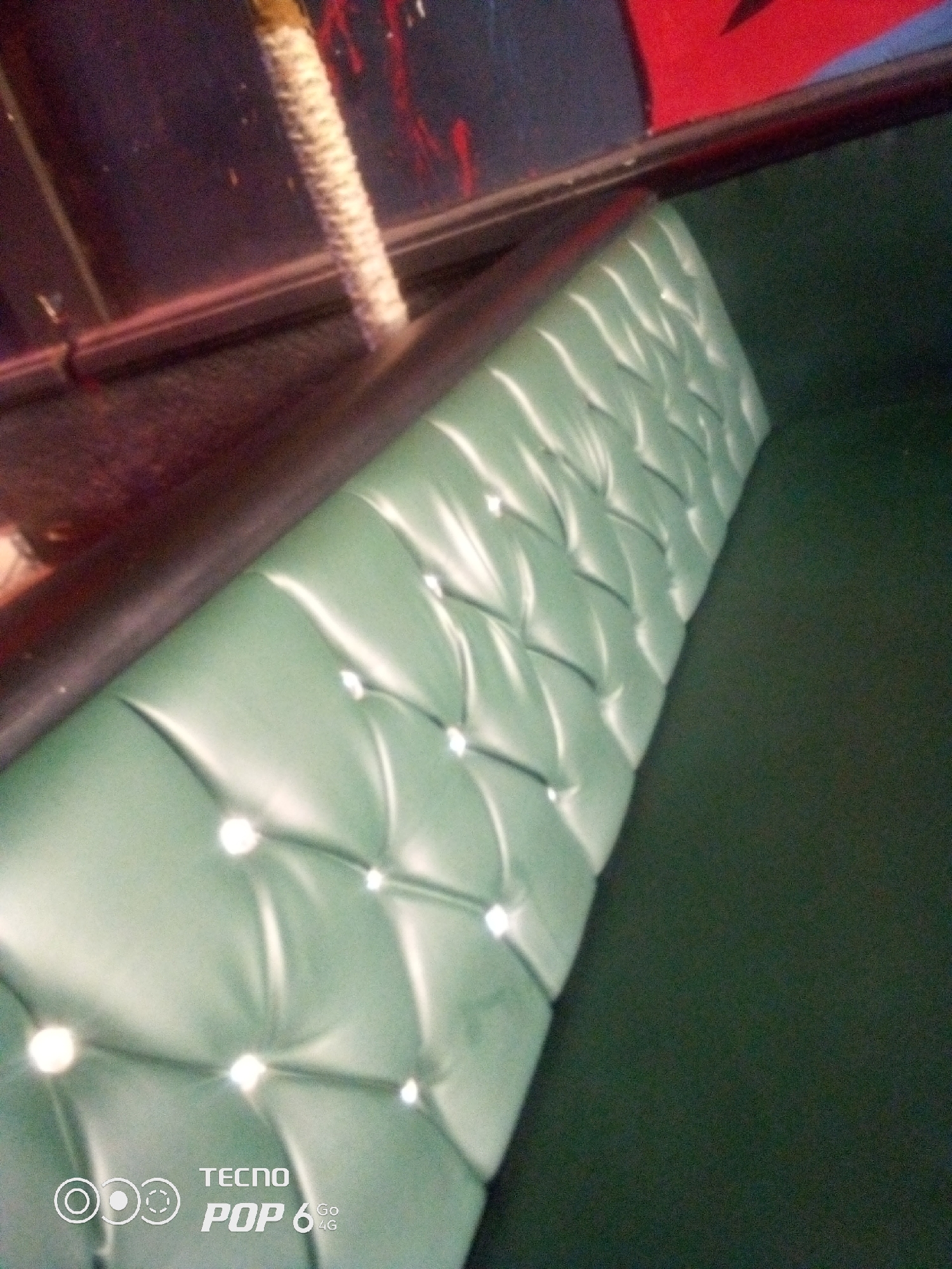 The image shows a close-up, angled view of a green, tufted, upholstered bench or sofa. The upholstery is made of a shiny material, possibly faux leather, and is adorned with clear buttons or studs that create a diamond pattern. The bench is positioned next to a dark, possibly wooden surface, which is partially visible at the top left of the frame. Above this dark surface, there are hints of red and dark blue in the background, suggesting a colorful wall or decoration. The lighting in the image creates highlights on the upholstery, emphasizing its texture and sheen. The lower right portion of the image shows a dark, solid surface, likely the continuation of the seating area or floor. The bottom center of the image displays a watermark with the text "TECNO POP 6 GO 4G". There is no indication of people or any specific location within Howayi, Nigeria.