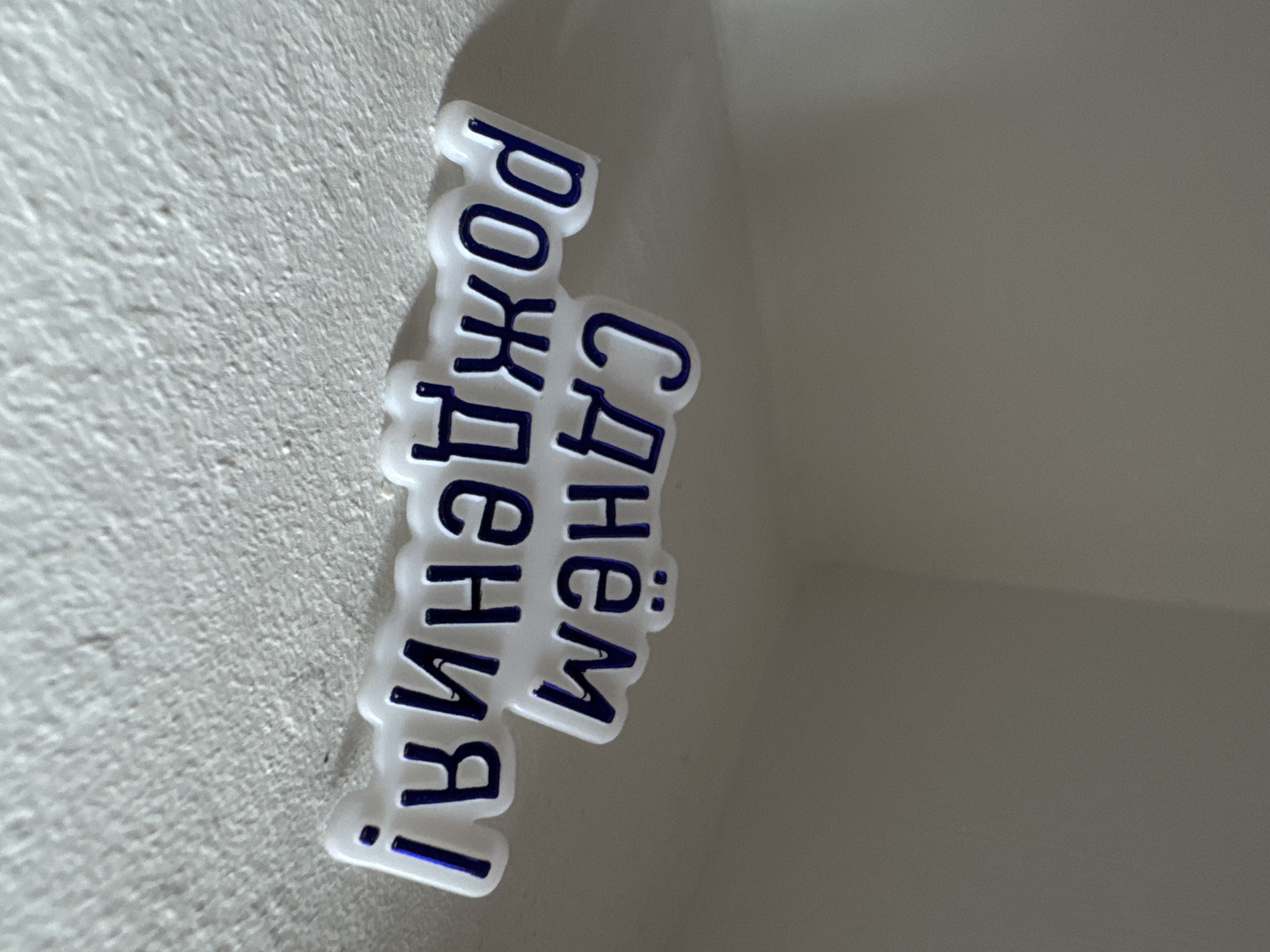 This image, captured in the city of Chaikovsky, Russia, depicts a white, irregularly shaped decorative object featuring blue Cyrillic text, resting on a textured, off-white surface. The text reads "С днём рождения!", translating to "Happy birthday!". The object, likely made of plastic or a similar rigid material, has raised blue letters with a slightly reflective quality, suggesting a glossy or metallic finish.
The textured surface beneath the object is coarse and granular, appearing to be a painted wall or similar architectural element, with visible variations in its relief. Shadows are cast from the object to its right and slightly behind it, indicating a light source originating from the upper left of the frame. The background consists of a smooth, lighter grey wall or interior surface, exhibiting a subtle transition from lighter tones on the left to slightly darker tones on the right, suggesting a corner or alcove. The focus is sharp on the foreground object and the textured surface, while the background appears softer. No people or other distinct subjects are visible in the scene.