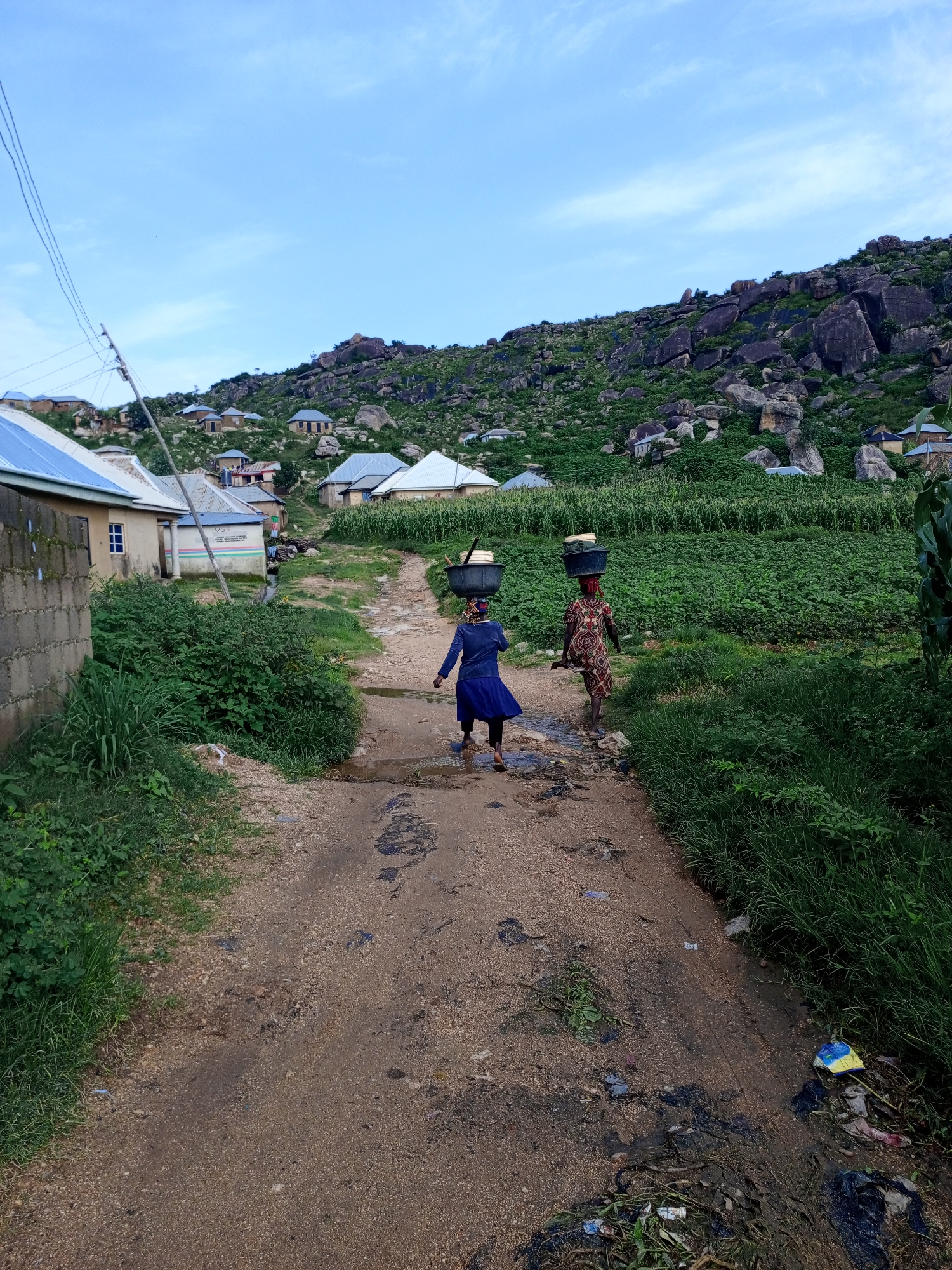 Two women are walking away from the camera down a dirt path in Rukuba Road, Nigeria. Both women are carrying large buckets of water on their heads. The woman in front is wearing a blue dress and the woman behind her is wearing a colorful patterned dress. The path is lined with lush green vegetation on either side. In the background, there are several houses and a rocky hillside covered in trees and shrubs. The sky is a clear blue with a few wispy clouds.