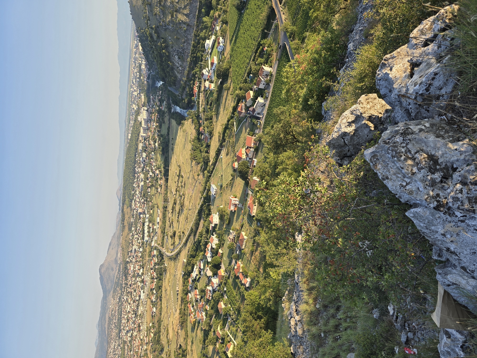 This is a high-angle, wide shot of a city situated in a valley with a river running through it. The city is densely populated with a mix of buildings, including residential homes with red roofs and larger, modern structures. Green fields and trees surround the city, with a highway visible as it winds through the landscape. In the foreground, a rocky, grassy hillside is present, with some vegetation and a small, brown object that may be a tarp or bag. The sky is clear and blue, and the lighting suggests it is late afternoon or early evening. The overall scene depicts a sprawling urban area integrated with a natural landscape under a clear sky.