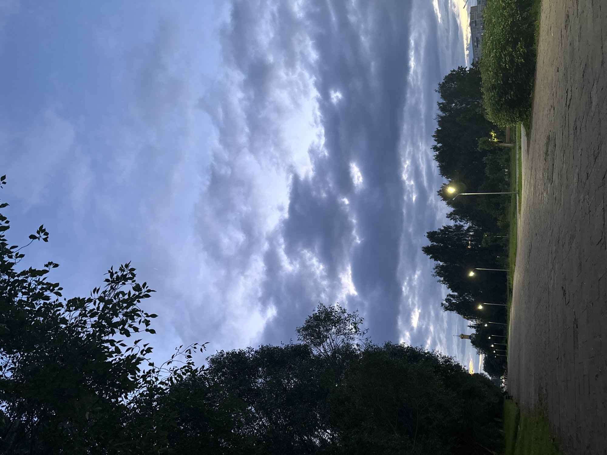 The image presents an outdoor scene at dusk or early night, depicting a park pathway or promenade in Naberezhnye Chelny, Russia. The sky occupies the upper portion, characterized by dark, heavy stratocumulus clouds interspersed with lighter blue patches and areas illuminated from behind, indicative of twilight. A faint, thin white line, possibly a contrail, is visible in the upper-left sky.
The left side of the frame is dominated by dense, dark green foliage from large trees, with specific leaves visible against the sky in the upper left. A wide, paved pathway, constructed from dark, irregular paving stones, extends from the foreground into the distance on the left.
Multiple street lamps with bright, yellowish-white lights are active, illuminating the pathway and surrounding elements. A distinct line of these lamps runs along the right side of the pathway, receding into the midground. Beyond the pathway, a continuous line of dark green trees forms a barrier. In the distant midground on the left, a golden onion dome and spire, consistent with the architecture of the Church of the Holy Ascension in Naberezhnye Chelny, are visible above the tree line.
In the foreground on the far right, a large, densely foliated green bush or hedge is brightly illuminated by an adjacent street lamp. Further back on the right, a multi-story building with a light-colored facade is partially visible above the tree line and bush. Several indistinct human figures are observable walking along the pathway in the far distance on the left. A park bench is faintly discernible near trees on the right side of the distant pathway. The overall scene conveys a publicly accessible recreational area with artificial illumination active against a fading natural light.