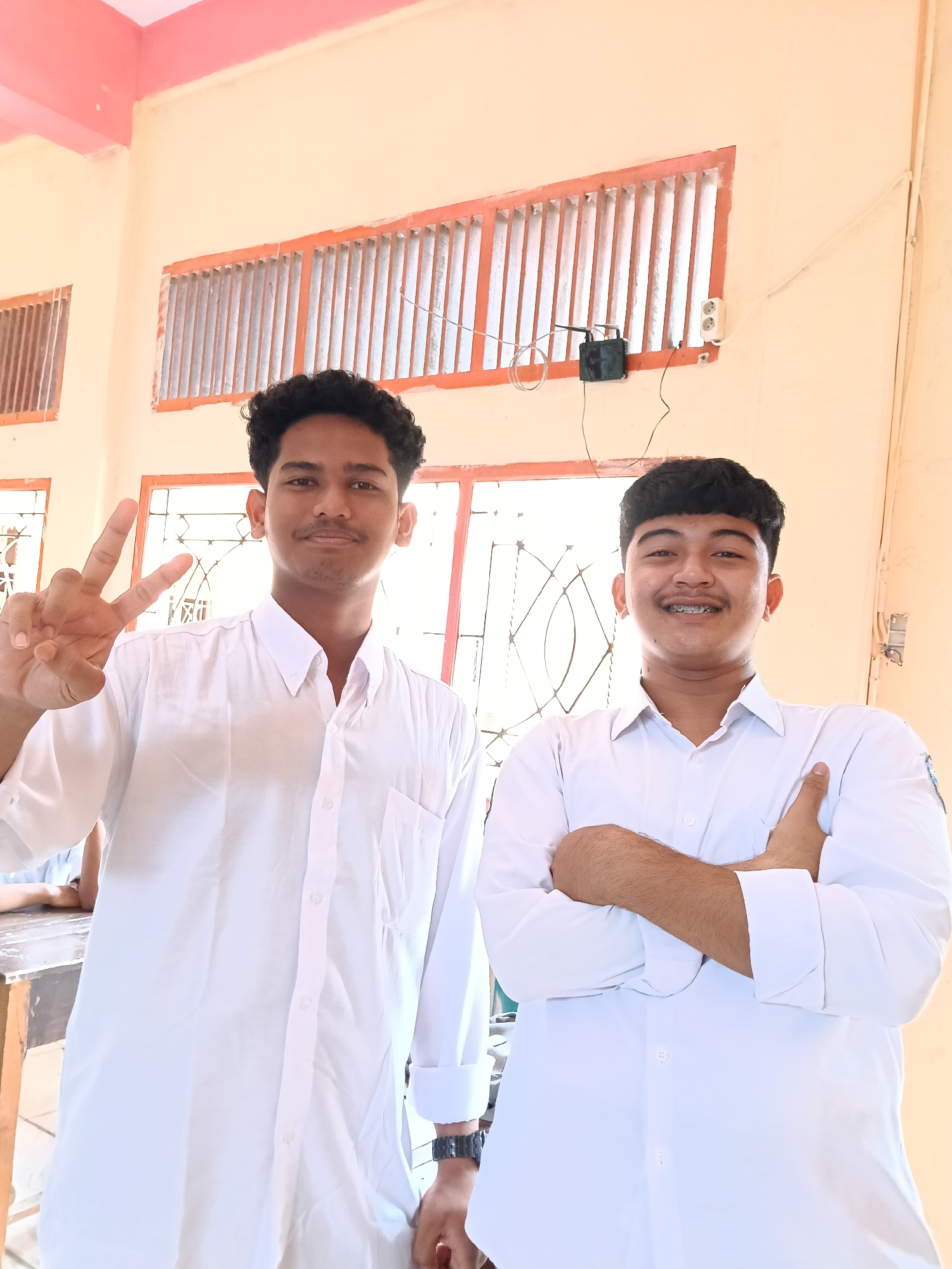 Two young men are standing indoors. The man on the left is holding up two fingers in a peace sign. The man on the right has his arms crossed. Both are wearing white shirts. Behind them is a window with bars, and a wall with an electrical outlet. The location appears to be in Lhokseumawe, Indonesia, based on visual indicators associated with the people in the image.