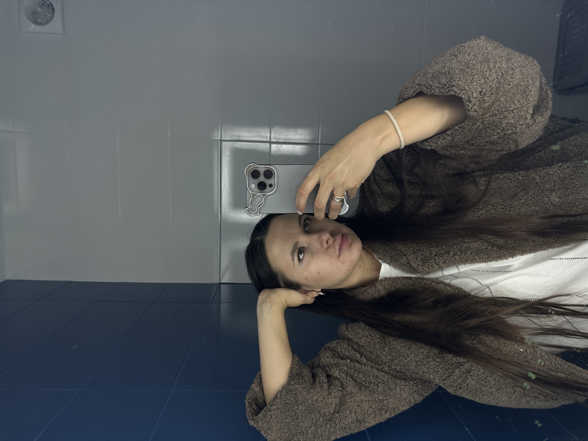 A young woman is lying on a navy blue tiled floor, wearing a brown fluffy jacket and a white t-shirt. She is holding a smartphone in her right hand, presumably taking a selfie. Her long brown hair is spread out on the floor. The background consists of white tiled walls, a vent, and a possible mirror reflection. This scene is possibly in a bathroom within a building in Akulovo (Gp Kubinka), Russia.