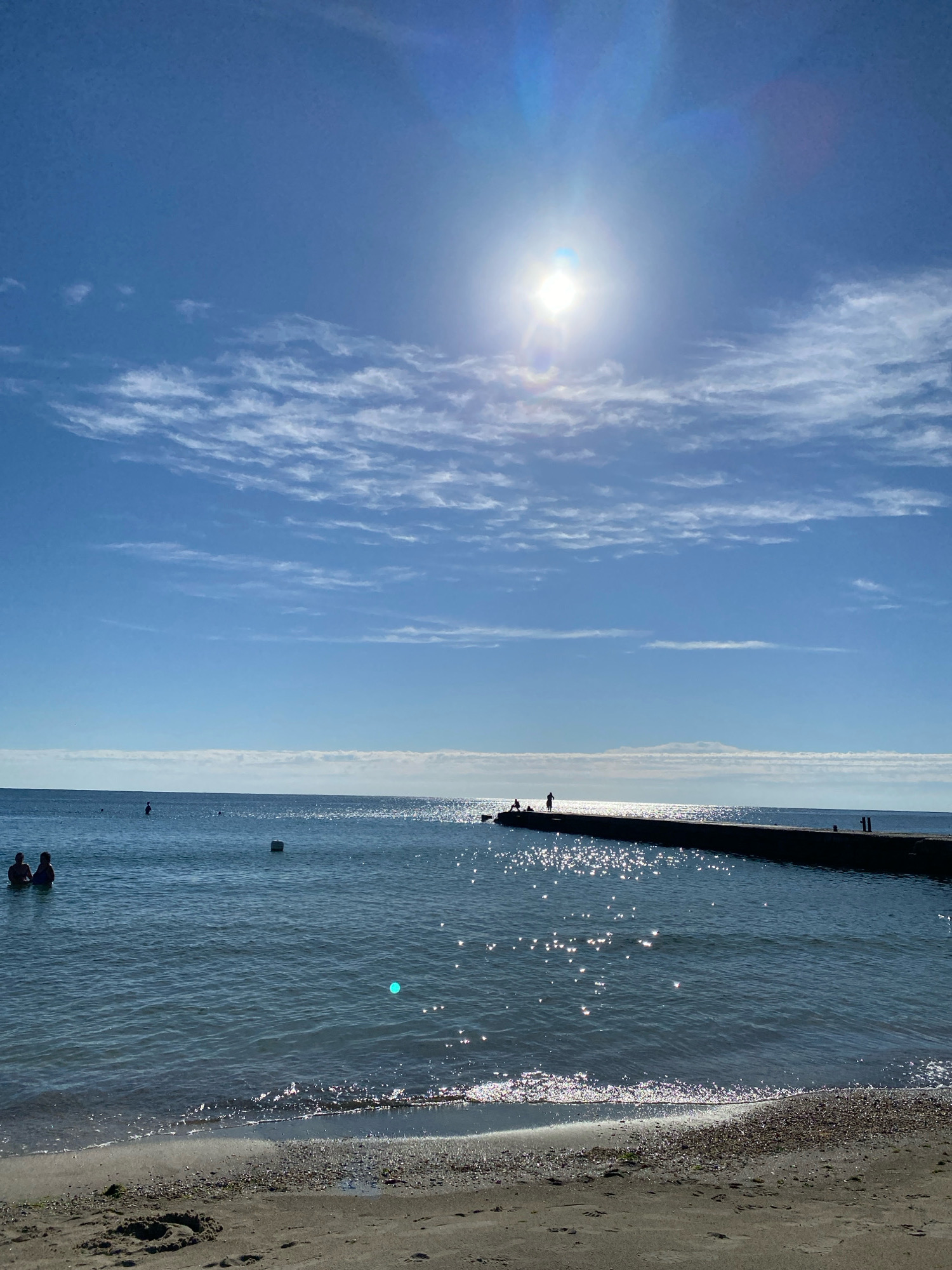 The image depicts a bright, sunny day at a beach in Odesa, Ukraine. The sun is high in the clear blue sky, with a few wispy clouds scattered across it. A long, dark pier extends into the calm, blue water. Several silhouetted figures are visible on the pier, some appearing to be standing and others sitting. In the distance, a few more figures are in the water, and there's a white buoy. The foreground shows a sandy beach with a few shells and footprints. The water is sparkling with sunlight.