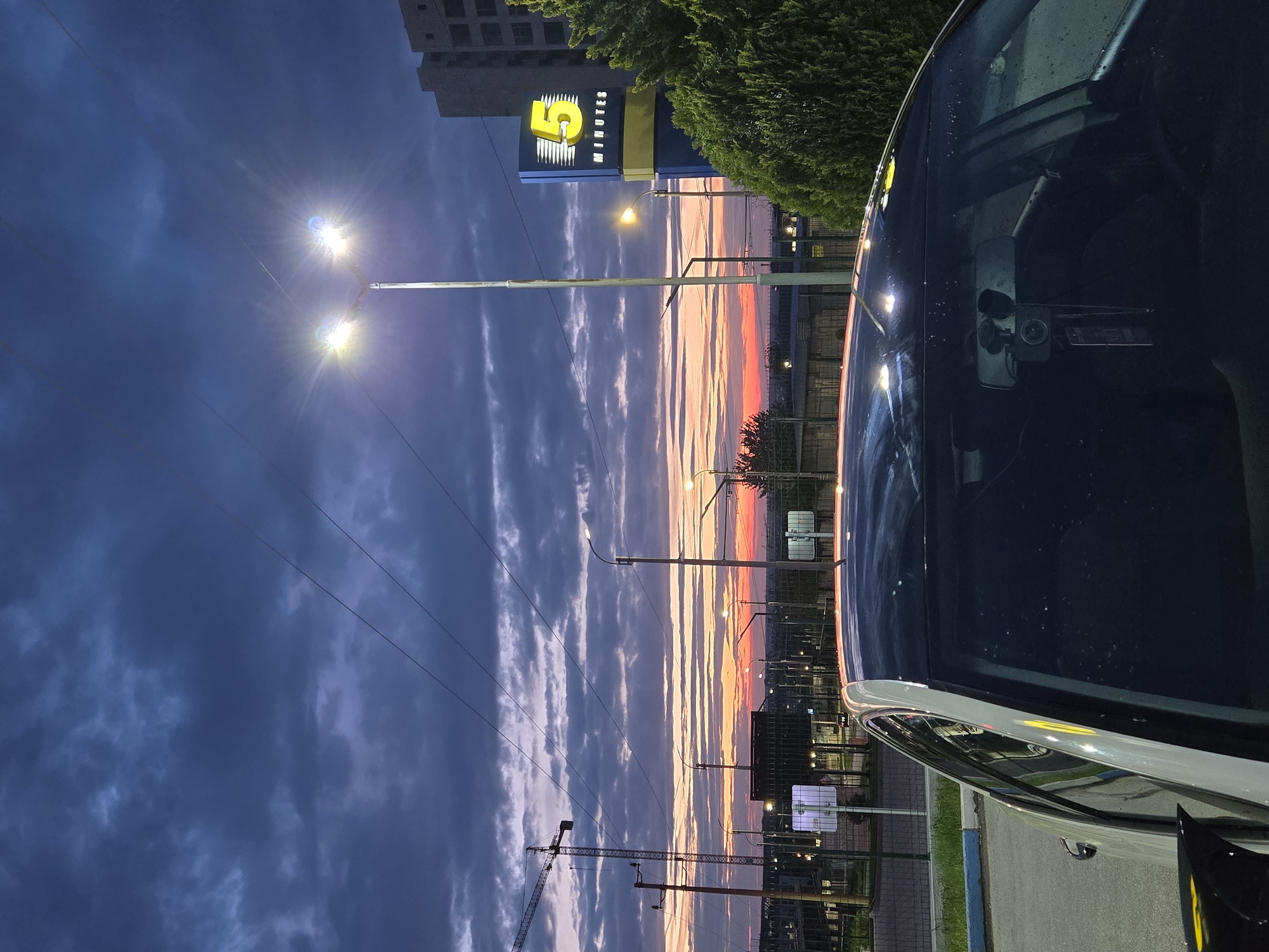 The image captures an urban twilight scene in Pleven, Bulgaria, likely at sunrise or sunset, given the vibrant colors of the sky and the illuminated artificial lights. The perspective is from inside or very close to a white car, a portion of which is visible on the right side of the frame, showing its sleek profile and reflections, along with water droplets on its surface.
The sky dominates the upper and left parts of the composition, transitioning from a deep, dusky blue with scattered dark clouds at the top to a dramatic horizon ablaze with hues of orange, pink, and pale yellow. Streaks of light-colored clouds extend across the blue sky, catching the ambient light.
Several streetlights are prominent, casting bright flares that pierce the semi-darkness. In the mid-ground, a tall sign with a bright yellow "5 MINUTES" logo stands out against the sky, flanked by dark green foliage. In the distance, an outline of a multi-story building and a construction crane are visible, along with power lines crisscrossing the frame. Other structures, more streetlights, and a blue painted curb are also present. The scene conveys a sense of calm and transition during the change of day.