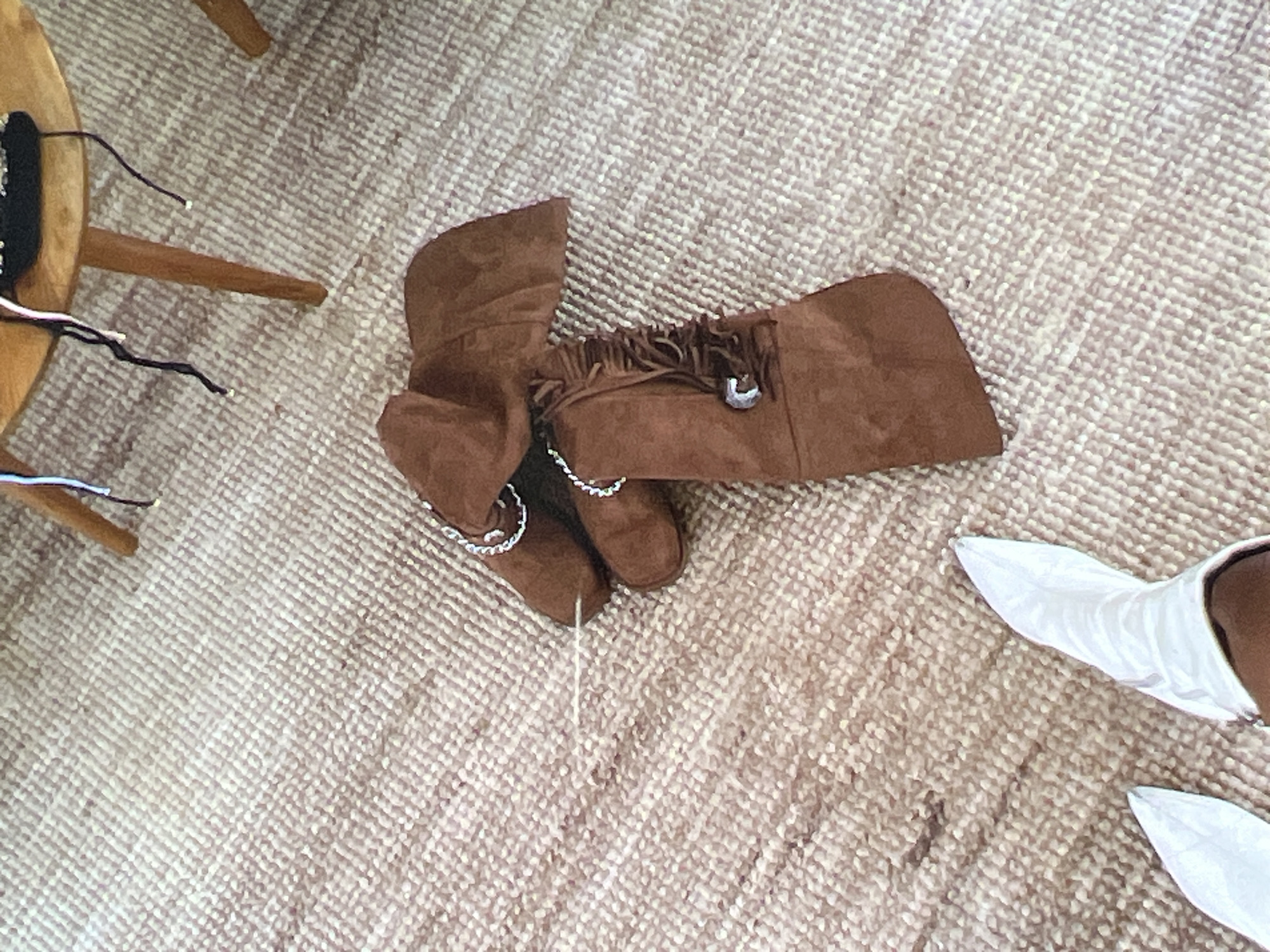 The image shows a top-down view of a light-colored, textured carpet. On the carpet, there is a pair of brown suede cowboy boots with fringe detailing and silver accents. Next to the cowboy boots, there are two white pointed-toe boots. In the background, part of a round, wooden table with dark strings hanging from it is visible. The scene appears to be indoors, with no indication of the specific location within Singapore.