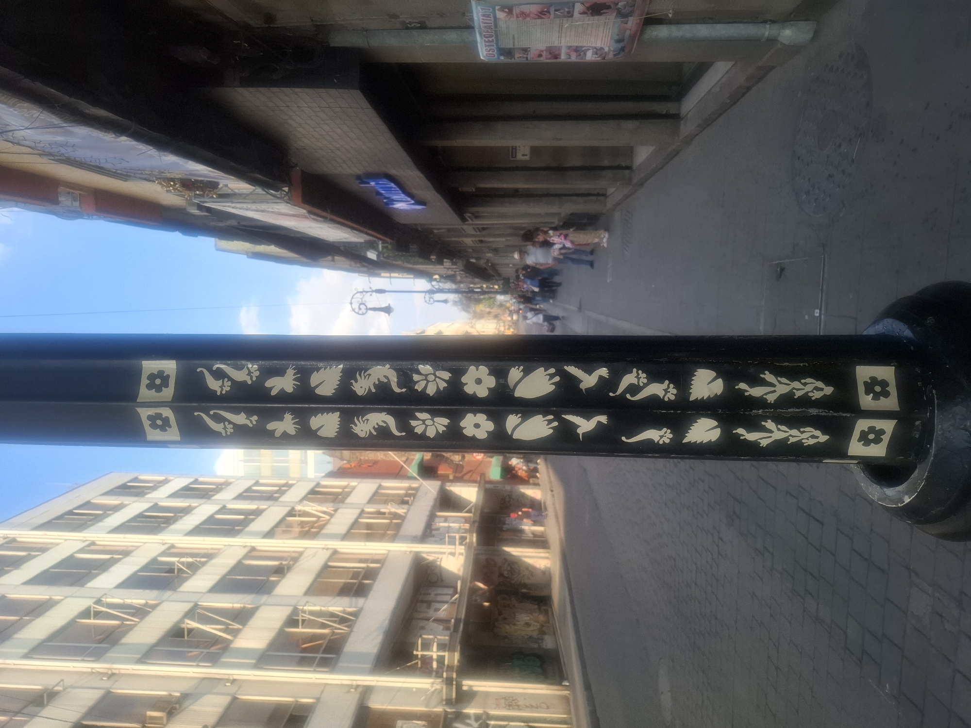 The image depicts a street scene in Mexico City, Mexico, viewed through a decorated lamppost. The lamppost is black with white floral and animal motifs. Behind the lamppost, on the left, is a multi-story building with a grid-like façade and scaffolding visible. The sky is blue and clear. On the right, a pedestrian street stretches into the distance, populated with people walking. Buildings line the street, and advertisements are visible on some of them. The street appears to be paved with cobblestones.