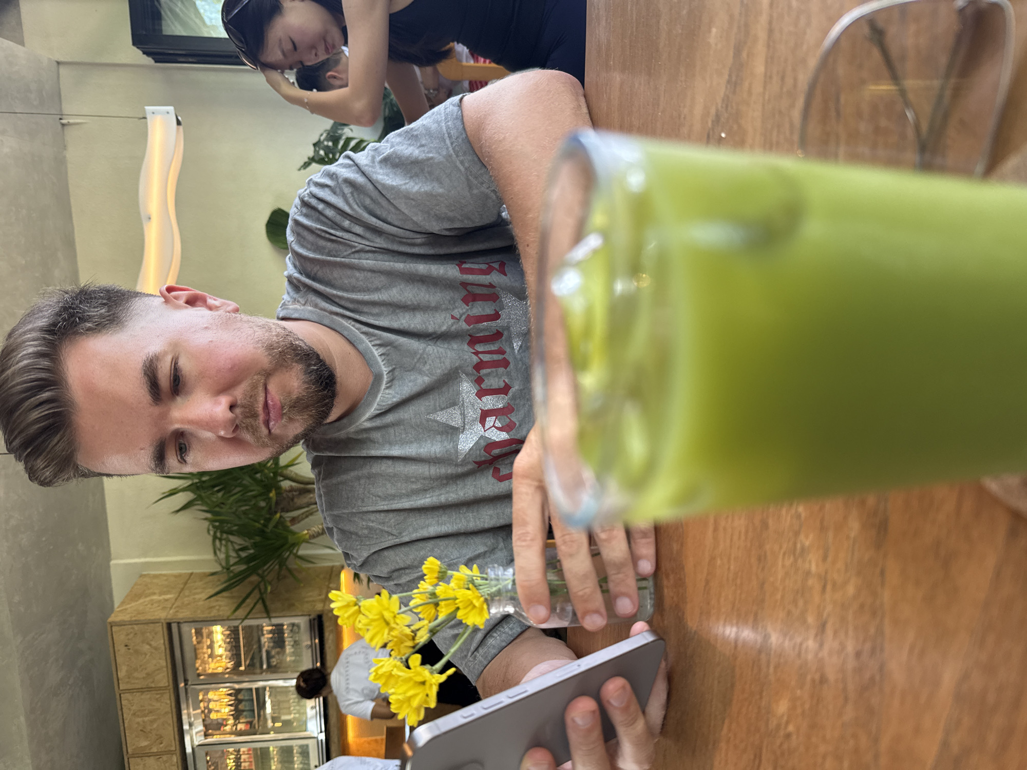 The image, taken indoors, shows a man sitting at a wooden table in what appears to be a restaurant. The table also holds a glass of green liquid, a small vase of yellow flowers, and a smartphone in his hand. The man is wearing a grey t-shirt. Behind him is another person with sunglasses on their head and a display case containing what looks like food or drinks. The location is identified as North Kuta, Indonesia.