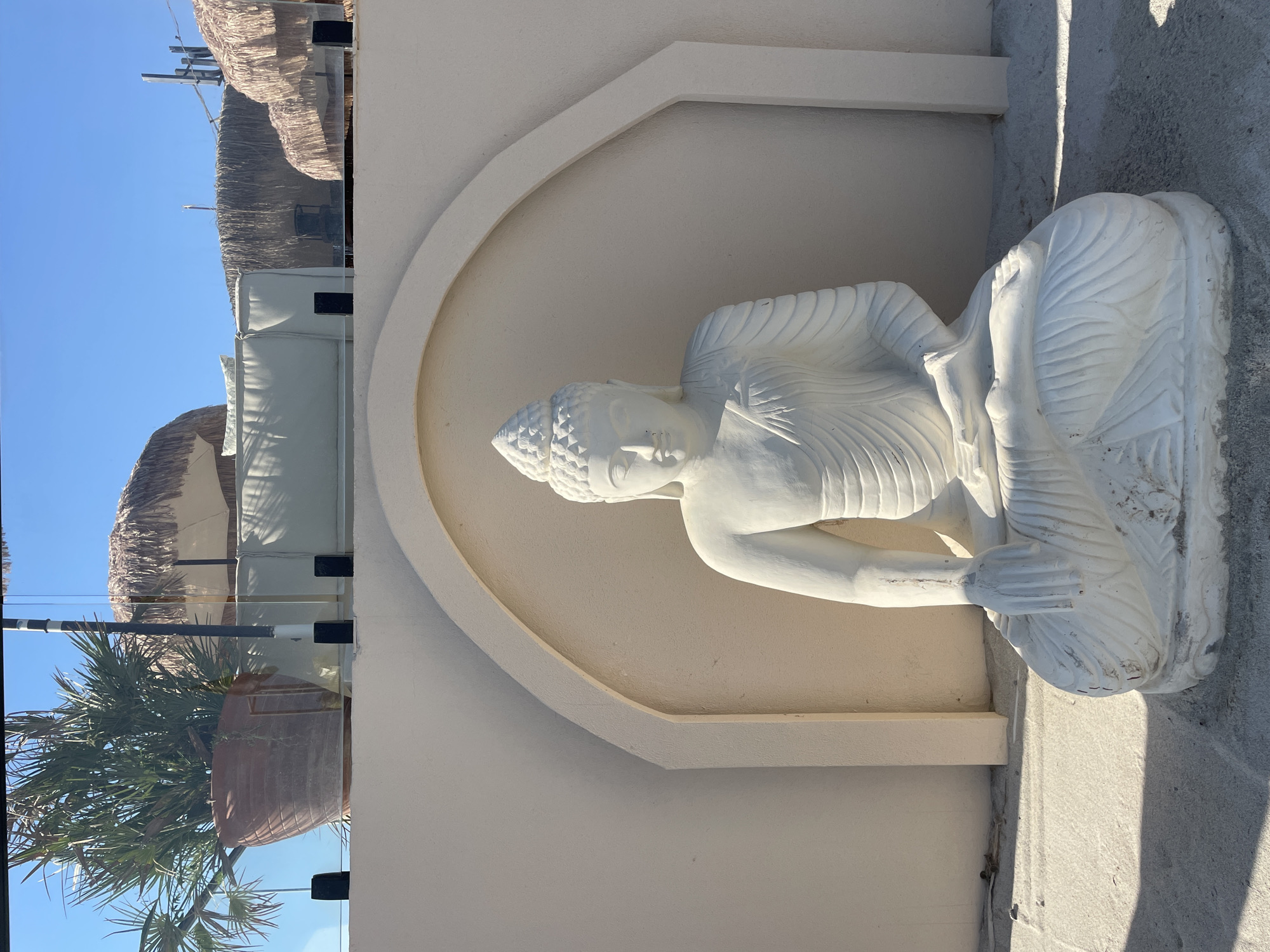 A white statue of Buddha is seated on a sandy surface in front of an arched alcove. The statue is facing forward with its legs crossed and hands resting in its lap. The Buddha has a serene expression on its face, with closed eyes and a subtle smile. Behind the statue, a cream-colored wall forms the alcove, with an arched opening. Above the wall, in the background, are elements of a resort or outdoor seating area, including thatched umbrellas, lounge chairs, and a potted palm tree, all under a clear blue sky. The scene appears to be outdoors, possibly in a sunny coastal location.