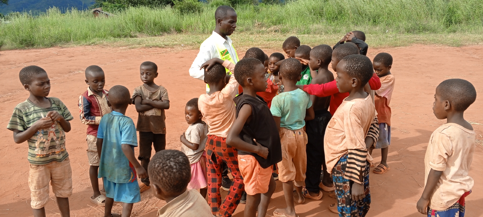 A group of approximately 20 children are gathered in an open, dirt area near Ngalula, Democratic Republic of the Congo. An adult male is standing among the children, placing his hand on the head of one of the children. The adult is wearing a white shirt and a yellow reflective vest. The children's clothing is varied. The background includes tall green grass and a building.