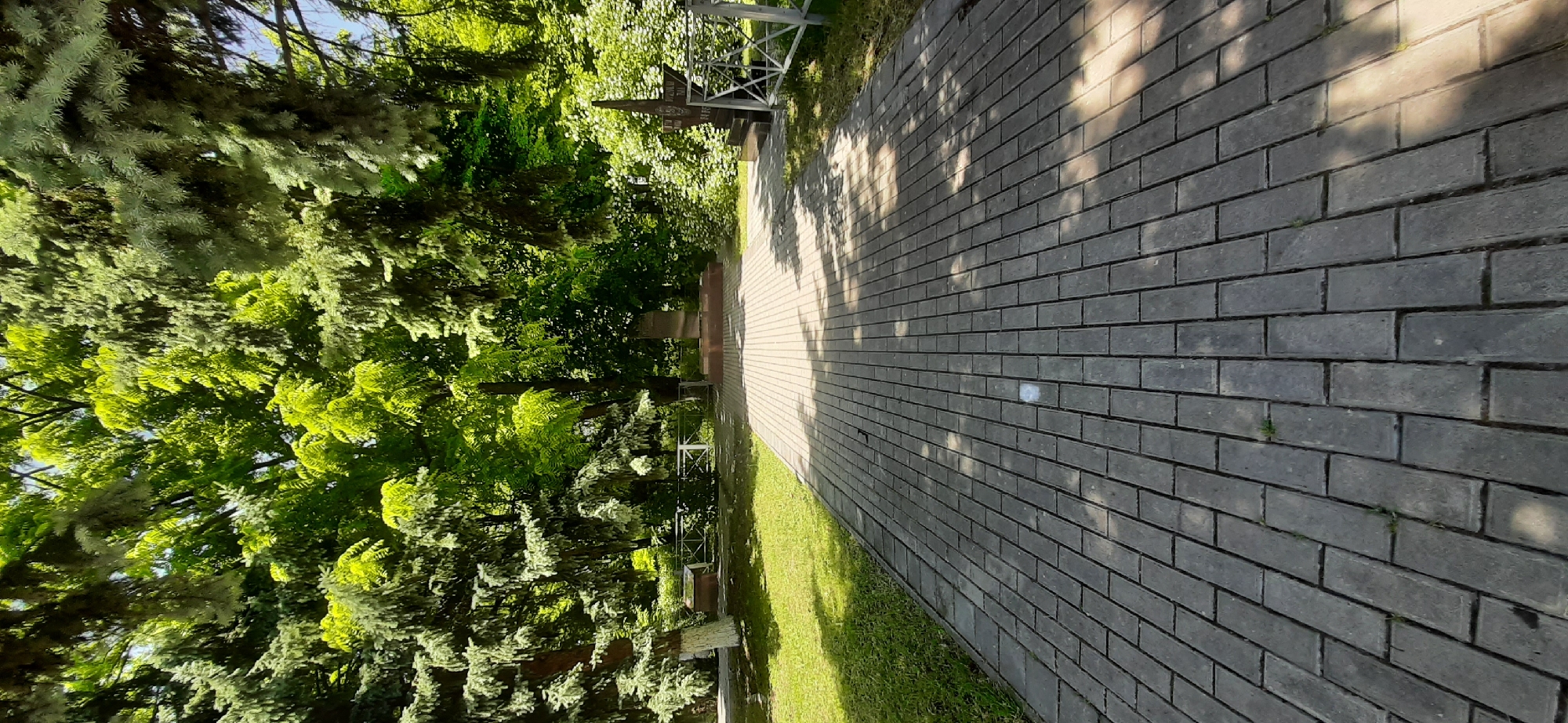 The image depicts a paved pathway leading through a park-like setting with lush green trees and grass. In the background, there are monuments and a white metal fence. The pathway is made of rectangular grey pavers and is partially covered in dappled sunlight and shadows from the surrounding trees. There are no people visible in the image. The scene appears to be a memorial or a quiet outdoor space within the city of Yessentukskaya, Russia.