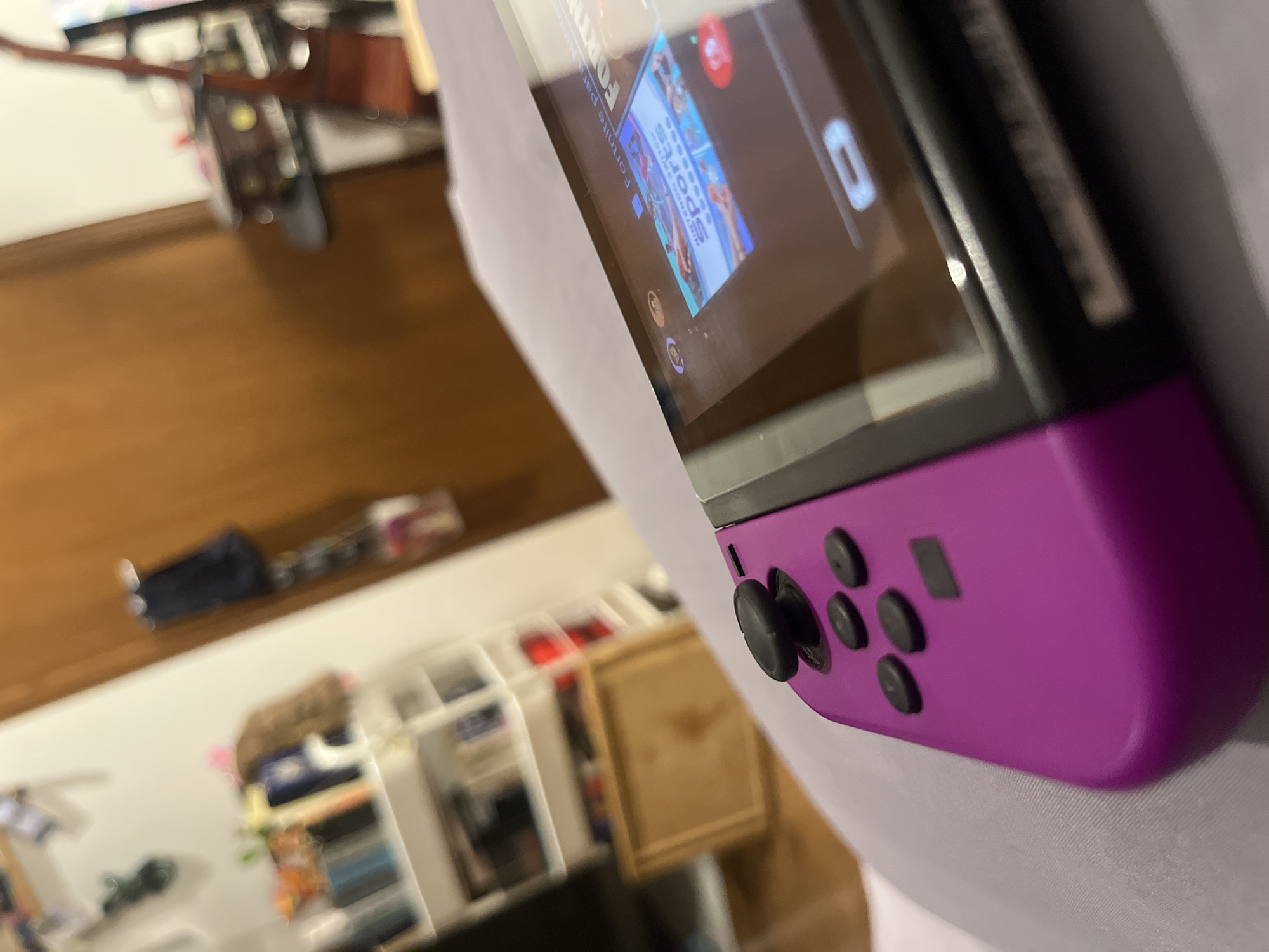 The image shows a close-up view of a Nintendo Switch console with a purple Joy-Con attached. The screen displays icons for Fortnite and Nintendo Switch Sports. In the blurred background, a bookshelf and a wooden cabinet are visible. The environment appears to be indoors, possibly a home setting. There is no information to determine that the image was taken in Mexico City, Mexico.