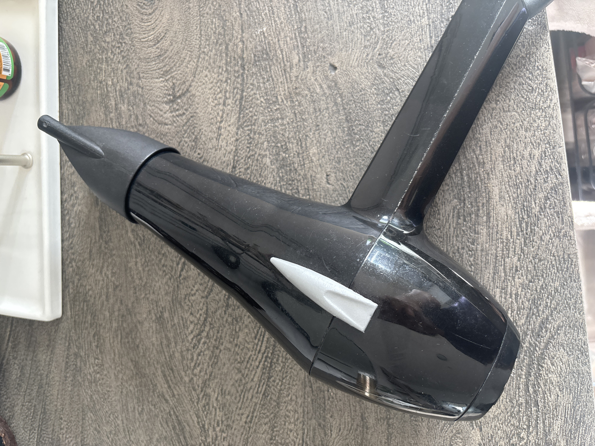 A black hairdryer is placed on a textured, dark gray surface, likely a table. The hairdryer has a silver accent on its body. In the background, a white tray and part of a bottle with a label are visible. The scene appears to be indoors, possibly a home or hotel room in Dubai, United Arab Emirates. There are no people or active interactions depicted. The lighting suggests an overhead or natural light source.