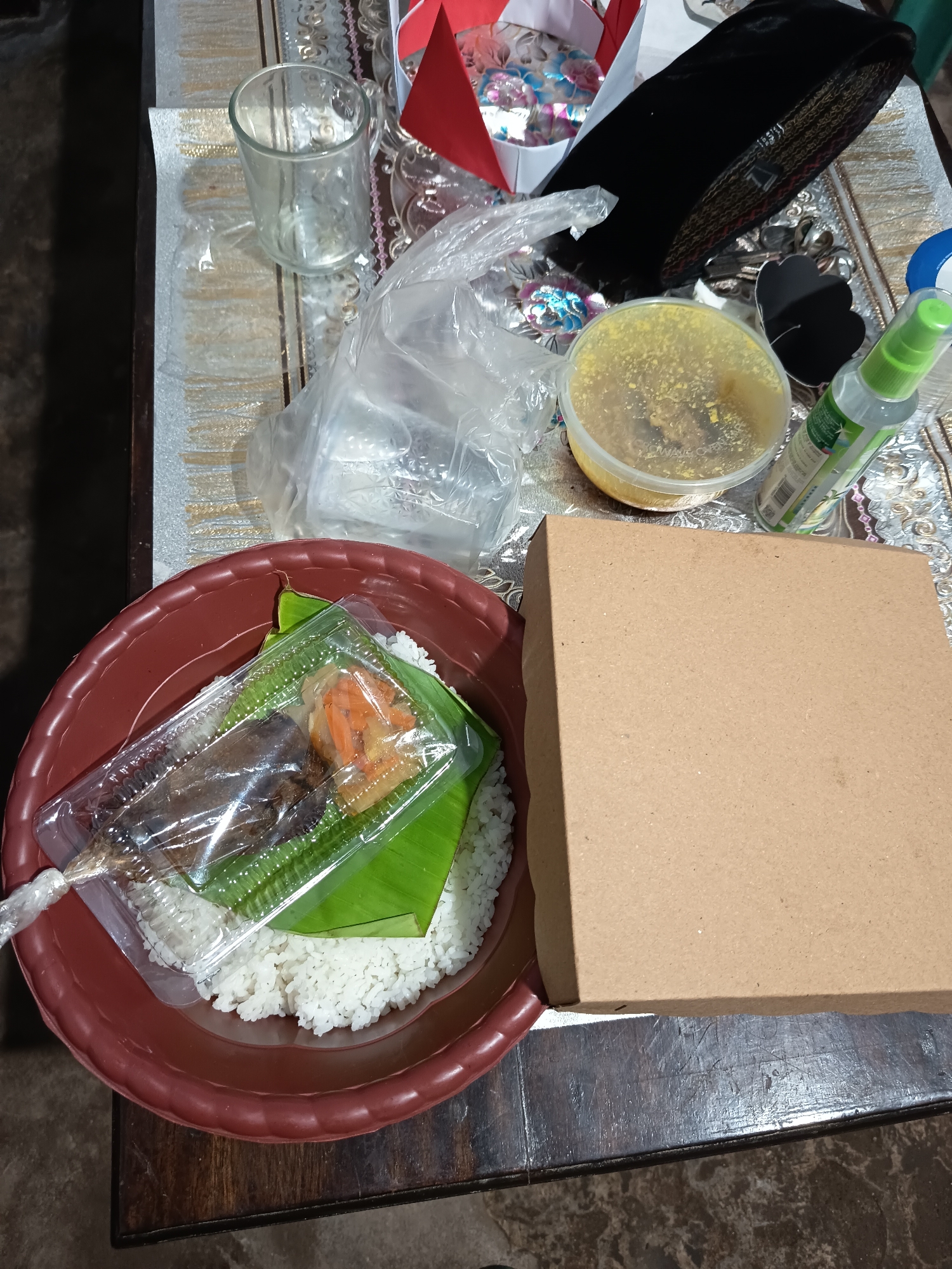 The image depicts a table setting, likely in Krampilan, Indonesia, with a meal and other items arranged on a patterned tablecloth. The central focus is a brown plastic bowl containing white rice, topped with a banana leaf. On top of the banana leaf is a clear plastic container with a piece of fish and some orange pickled vegetables. To the right of the bowl is a closed brown cardboard box. In the background, there are various objects including a clear glass, a plastic bag filled with items, a round plastic container with yellow contents, a black peci (traditional cap), and a green spray bottle. The lighting suggests an indoor setting, possibly a home or a small eatery.