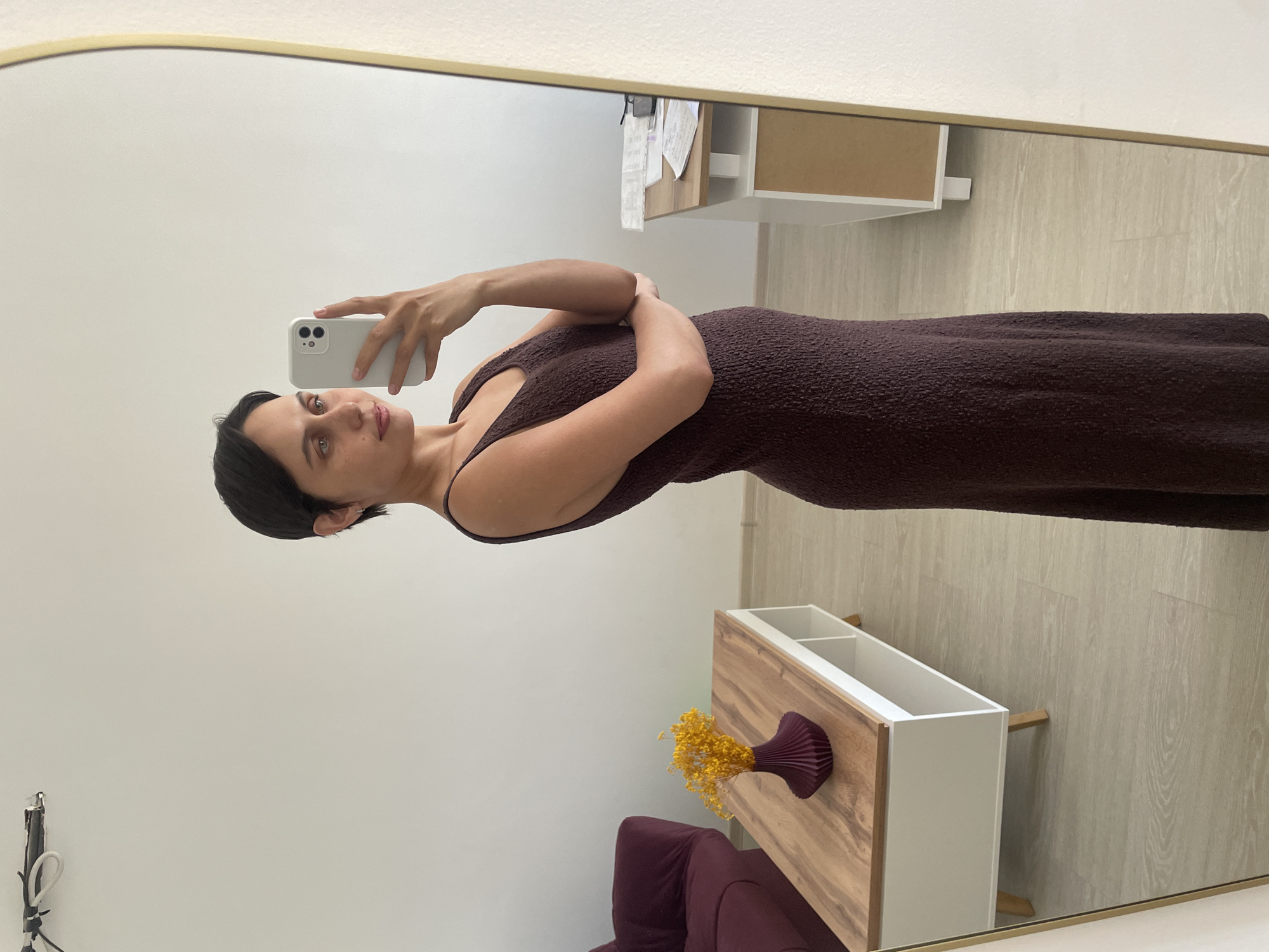 A woman is taking a selfie in a mirror. She has short dark hair and is wearing a dark brown dress. She's holding a white smartphone. The background shows a room with white walls, light-colored flooring, a wooden and white cabinet with a vase of yellow flowers on it, and a dark purple armchair. Papers are stuck on an upper white cabinet. The image appears to be taken indoors. There is no information to suggest the location is in Izhevsk, Russia.
