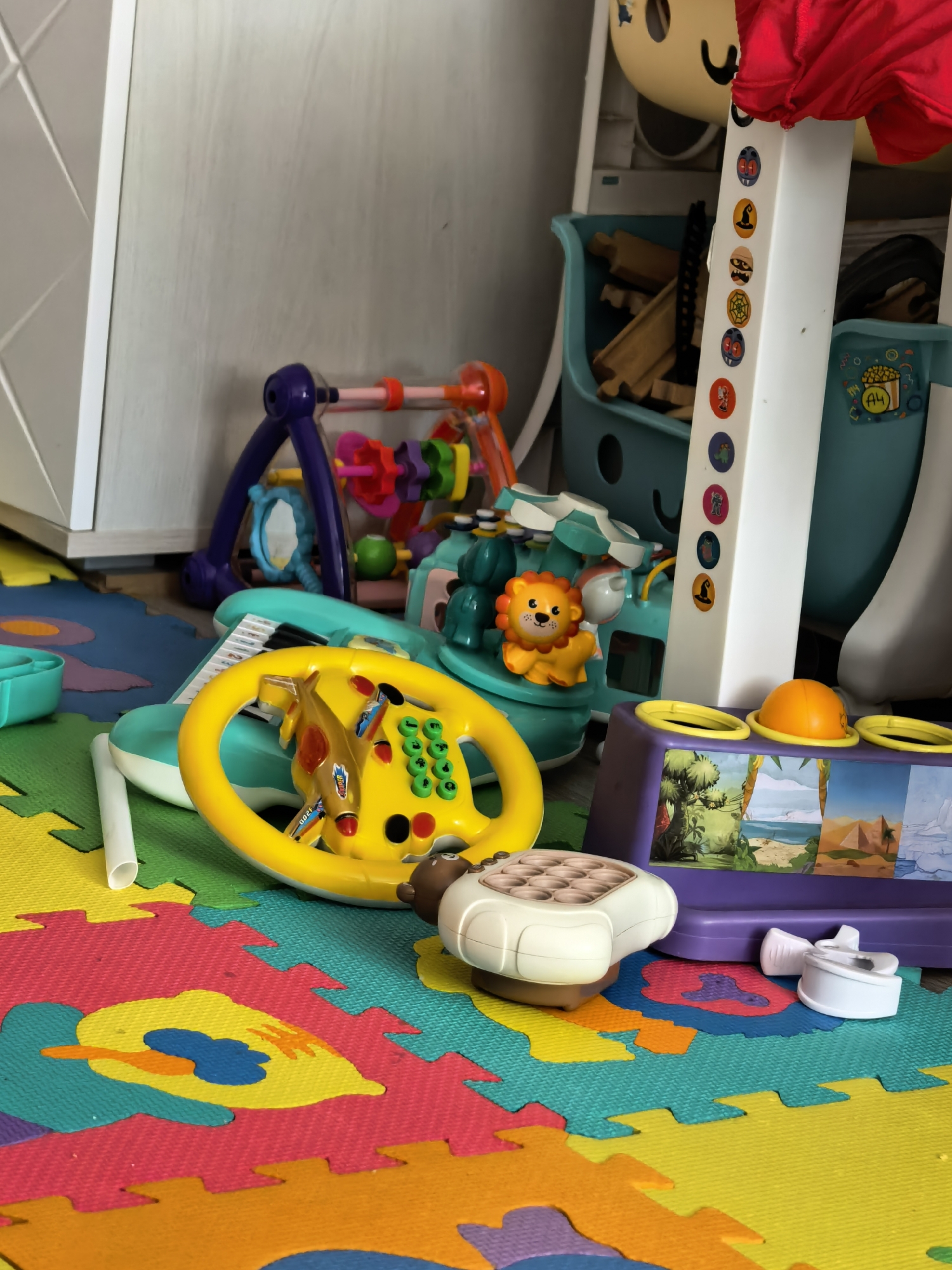 The image depicts a collection of children's toys scattered on colorful interlocking foam mats. The toys include a yellow steering wheel with buttons, a plastic lion toy on a light green base, a purple baby gym, a purple toy with illustrated panels, a toy phone, and a white tower with Halloween-themed stickers. A blue toy storage bin with wooden train tracks inside is visible in the background. The location is unspecified in the image. Sterlitamak, Russia is not evident from this image.