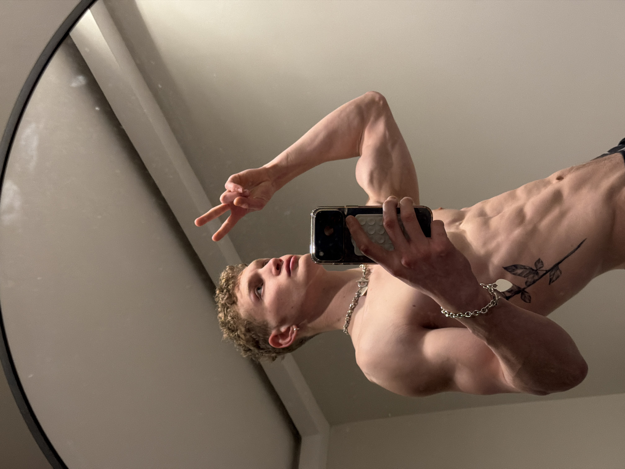 A young, shirtless male with short, curly blonde hair is depicted in a mirror selfie. He holds a dark-cased smartphone in his right hand, positioned to capture his reflection. His left arm is raised, with his hand forming a V-sign. The subject exhibits a muscular physique, with visible abdominal muscles, biceps, and shoulders. He wears a silver-colored chain necklace with a T-bar clasp and a silver-colored link bracelet featuring a heart-shaped charm on his left wrist. A tattoo of a rose stem with leaves is present on his left lower ribcage/abdomen area. He is gazing upwards and to his right. The background reflected in the mirror consists of a plain, light grey-beige wall and a ceiling line. The mirror itself has a dark, rounded frame and shows minor surface smudges. The scene is an indoor setting; no specific location within the city of London, United Kingdom, can be determined from the visual information provided.