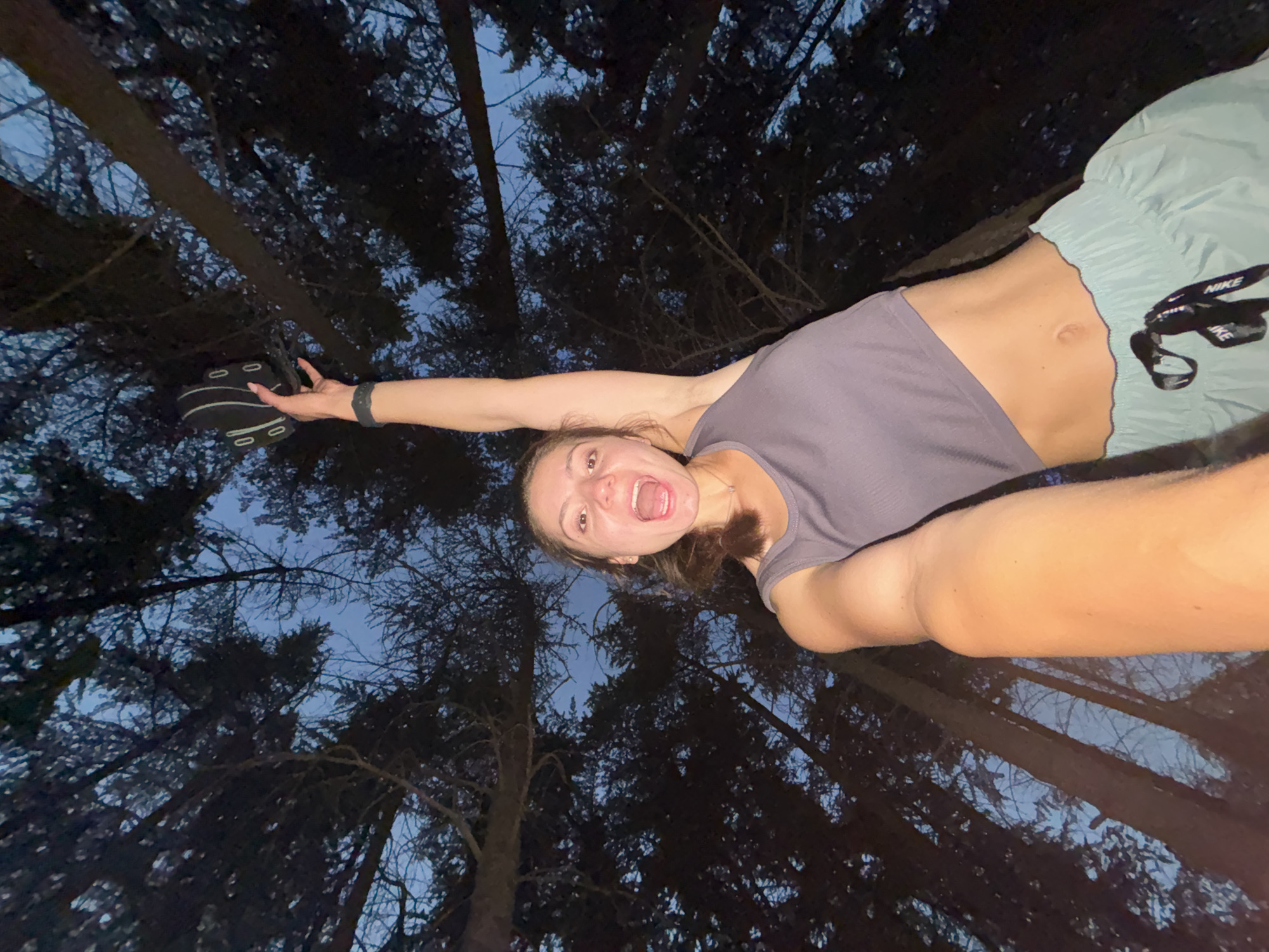 A young woman is captured in a low-angle, wide shot as she holds a black and white object above her head amidst the trees. The woman is wearing a gray crop top and light blue athletic shorts, and she is looking up towards the camera with her mouth open in an excited expression. Her right arm is extended upward, holding the object, which appears to be a tambourine. The background consists of dark trees against a twilight sky, suggesting an outdoor setting possibly in a forested area of Varna, Bulgaria, at dusk or dawn. The low angle of the shot emphasizes the height of the trees and creates a sense of dynamic energy from the woman's pose and expression.
