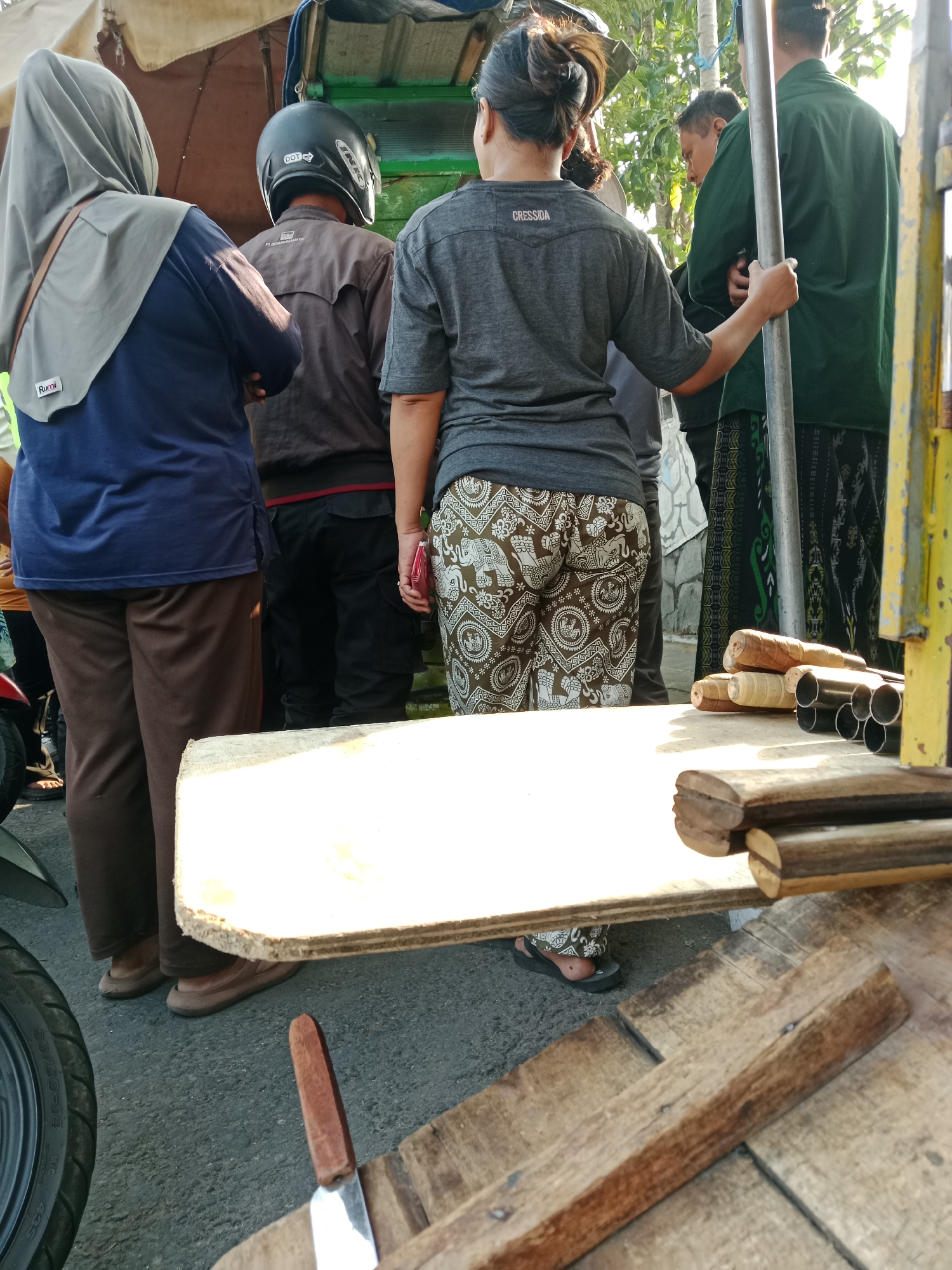In Kediri City, Indonesia, a street scene unfolds with individuals gathered near a market stall. In the foreground, a woman, facing away from the camera, wears a grey t-shirt with the word "CRESSIDA" visible on the back and patterned brown and white pants. To her left, another person, also facing away, wears a navy blue long-sleeved shirt and brown pants, with a grey hijab. Behind them, a person in a brown jacket and black pants, wearing a black helmet, stands with their back to the camera. To the right, a person in a dark green shirt and patterned pants stands near a metal pole. The stall features a light-colored wooden surface displaying several cylindrical wooden objects and metal tubes. A knife with a wooden handle is also visible on a wooden plank in the foreground. The background suggests an outdoor setting with sunlight casting shadows.