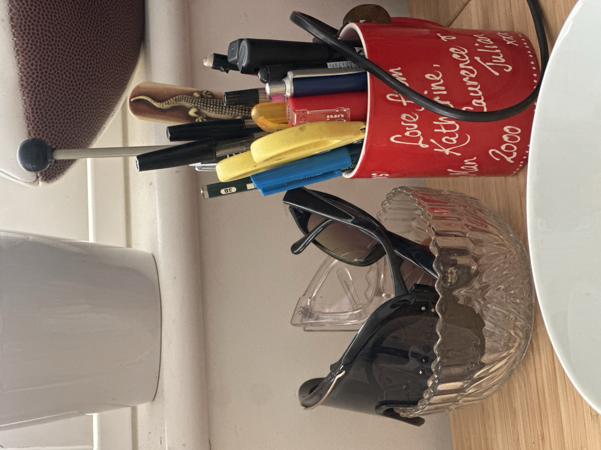 The image presents a still life scene of various personal items organized on a light-colored wooden surface, likely a desk or shelf in an indoor setting.
In the center, a vibrant red mug stands filled with an assortment of writing instruments and tools. These include multiple pens and markers in black, yellow, blue, and red, along with a pencil marked "3B" and what appears to be a stylus with a grey tip. A small, decorative item resembling a carved lizard or gecko is nestled among the pens. The mug itself features handwritten white text that reads: "Love from Man, Katherine, Laurence & Julian 2000 xxxx". A black cord or wire runs horizontally across the upper part of the mug.
Adjacent to the red mug, to its right, a clear, textured glass bowl holds several pairs of sunglasses, predominantly a dark-framed pair. Further right, a white ceramic or plastic plate or bowl is partially visible, resting on the wooden surface. To the left of the mug, a large, rounded object with a distinct brown, pebbled texture, indicative of a football or rugby ball, is partially seen. A tall white ceramic or plastic container is also present on the far left.
The items suggest a domestic environment, possibly in a home located in Wealden, United Kingdom. The lighting appears to be soft and ambient, suggesting daytime without direct harsh sunlight. No specific activity is depicted, making it a static arrangement of everyday objects. Other visible text includes "UNI" on a blue pen and a partial barcode on another writing tool.