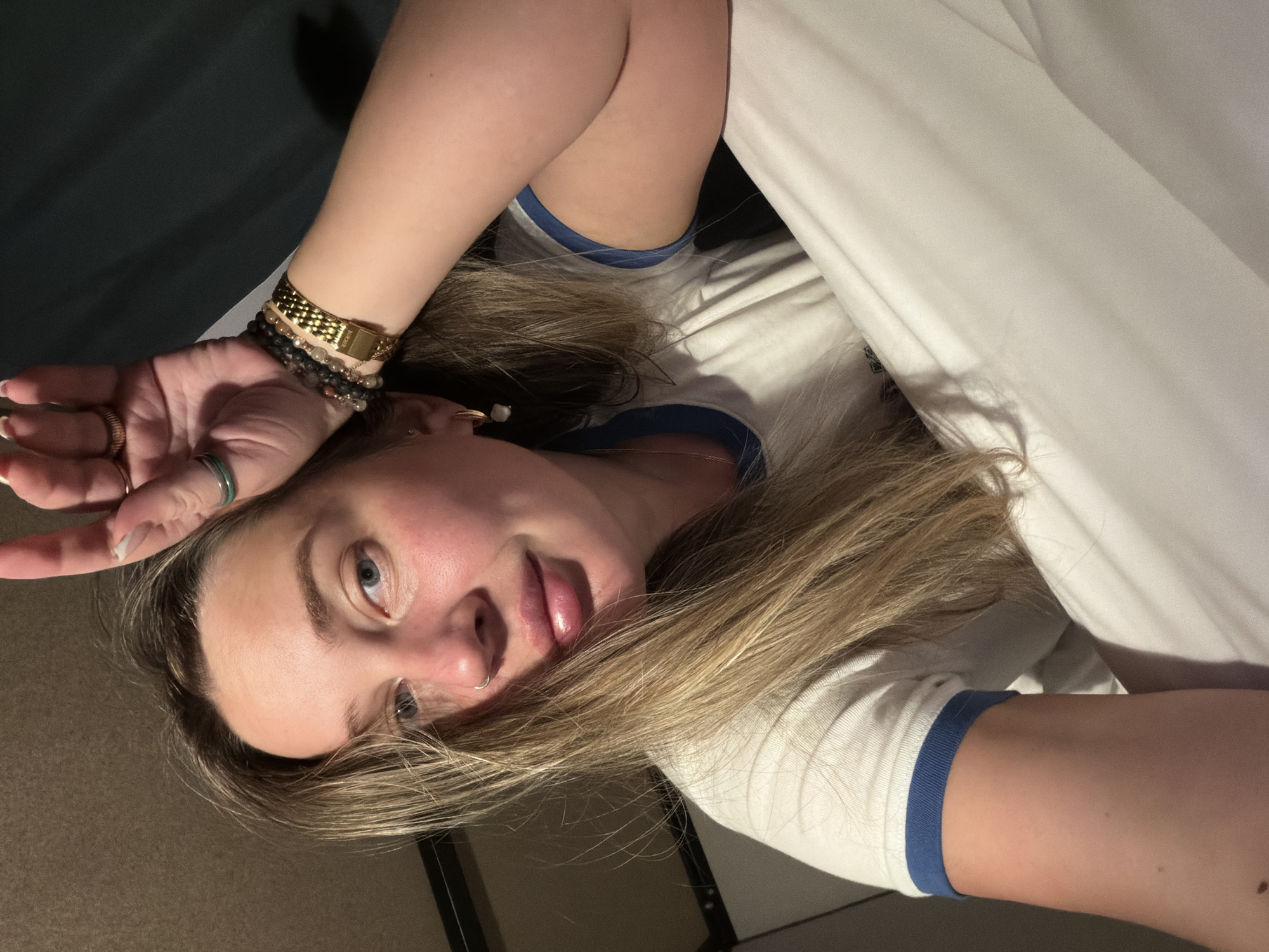 A young woman lies on a bed. She is wearing a white T-shirt with blue trim and several bracelets. She is indoors on the bed with white sheets. There is no indication that the image was taken in Moscow, Russia.