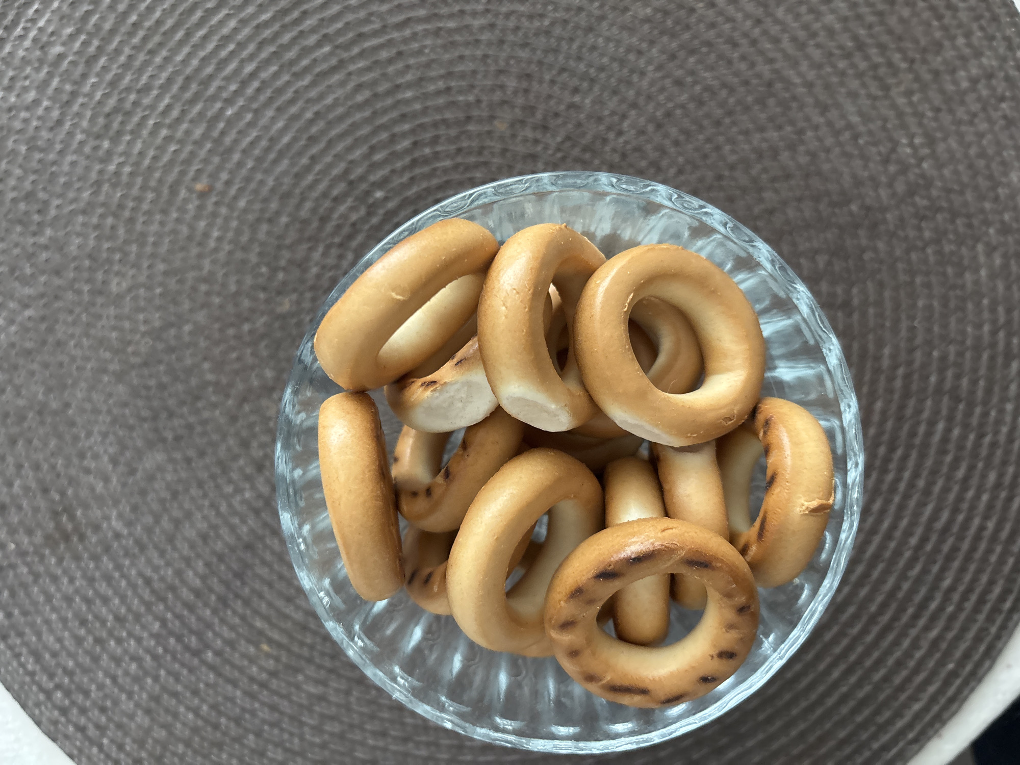 The image shows a clear glass bowl filled with small, golden-brown, ring-shaped pastries, likely bagels or "sushki". The bowl sits on a textured, brown surface, possibly a placemat. No people are visible. The location within Saint Petersburg, Russia, is not determinable from the image content.