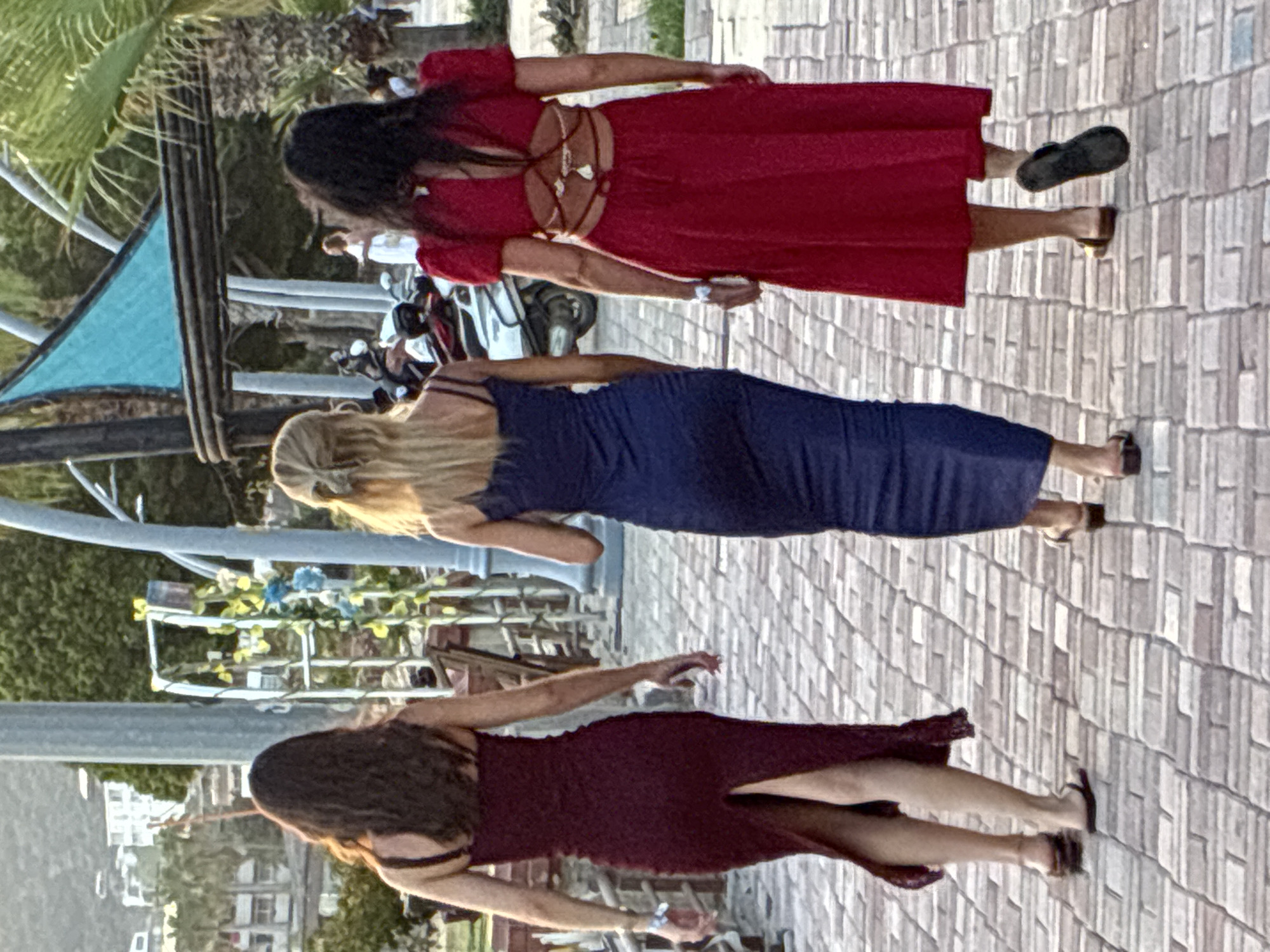 Three women are walking away from the camera on a paved walkway in Alanya, Turkey. The woman on the left is wearing a burgundy, form-fitting, floor-length dress with a slit up the back. She has dark, wavy hair. The woman in the middle is wearing a navy blue, form-fitting, floor-length dress and has blonde hair pulled back in a bun. The woman on the right is wearing a bright red, floor-length dress with short puffy sleeves and an open back with criss-cross straps. She has dark hair. In the background, there are palm trees, a building on a hill, a light blue awning supported by dark wood beams, and several motorcycles. The walkway is made of brick-like pavers.