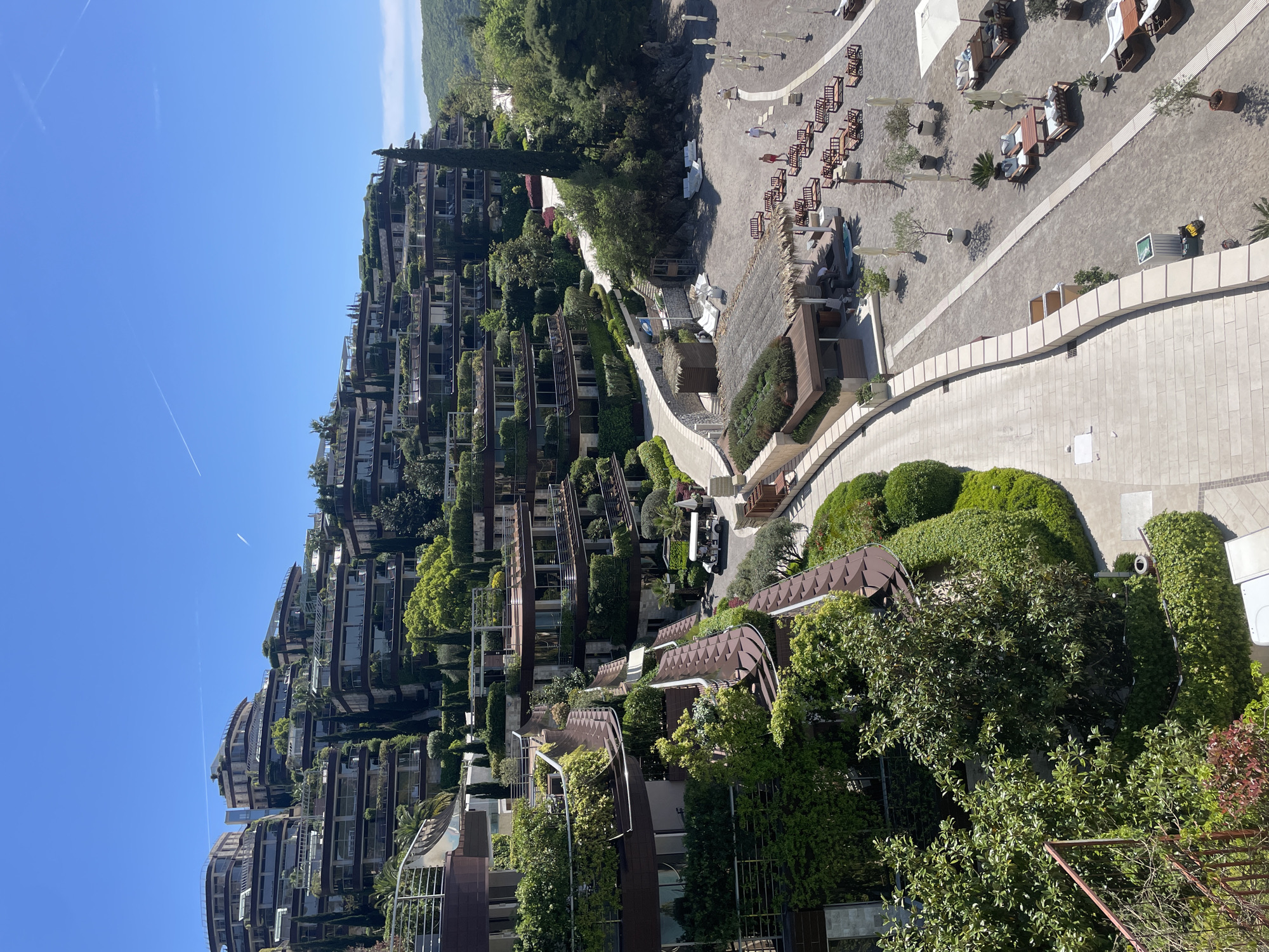The image captures an expansive, luxurious resort complex situated on a verdant hillside in Budva, Montenegro, under a clear, bright blue sky. The scene is dominated by multi-story, modern buildings with dark facades and extensive glass windows and balconies, arranged in a terraced, cascading design that blends into the natural slope. These structures are heavily integrated with lush green vegetation, including numerous trees, shrubs, and climbing plants that adorn balconies, rooftops, and pathways, creating a distinctive "green architecture" aesthetic.
A light-colored paved pathway winds through the resort, leading down to a wide, sandy or gravel-covered beach area. On this beach, numerous wooden sun loungers with white cushions, small tables, and white umbrellas are neatly arranged, suggesting a designated leisure zone. A few individuals are visible across the beach and paths; some appear to be relaxing on the loungers, while others are walking. A white golf cart is parked on one of the resort's pathways, indicating internal transport.
In the background, verdant hills and mountains rise, completing the scenic landscape. The sky is clear and bright, featuring faint white contrails from distant aircraft. The strong sunlight casts distinct shadows, indicating a time of day between late morning and early afternoon. The overall scene conveys a tranquil, luxurious resort environment designed for relaxation and harmony with nature. No visible text is clearly discernible.