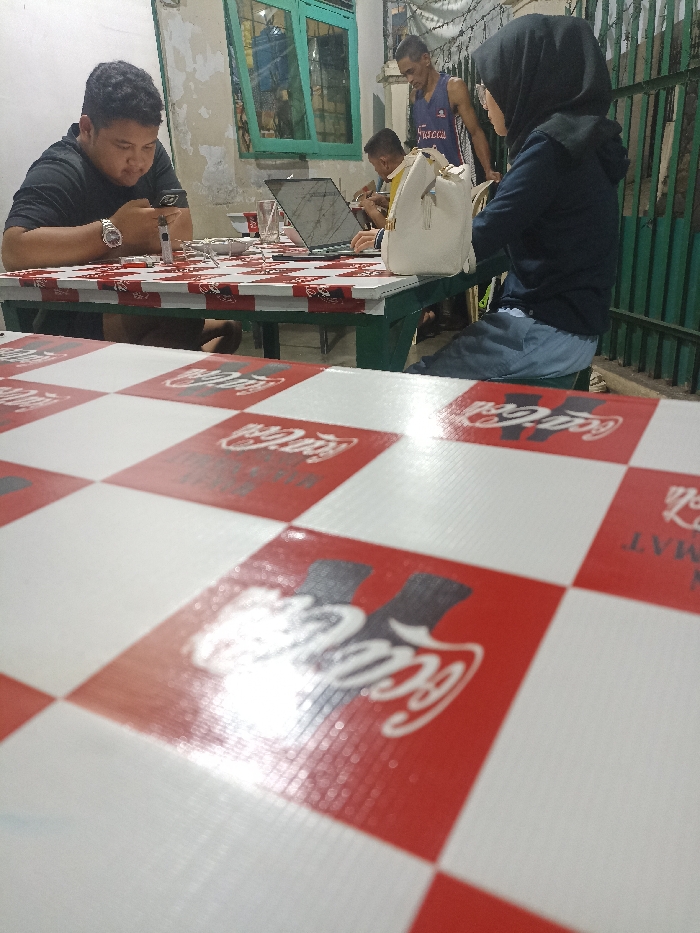 The image shows a partially obscured view of a checkered red and white table with a Coca-Cola logo in the foreground. The table is in front of a partially visible table with four chairs. One person sits at the table in the foreground, a young man in a black shirt, looking down at a smartphone. Two other people are partially visible in the background, both seated at the table in the foreground. One person is a young man in a blue shirt, looking at a laptop. The other person is a young woman wearing a long black jacket and headscarf, looking at a phone. There is a white purse next to the man in the blue shirt. The scene takes place in an outdoor cafe in Jatinangor, Indonesia, at coordinates -6.934 latitude, 107.778 longitude. There are green metal bars to the right side of the image, partially obscured by foliage.