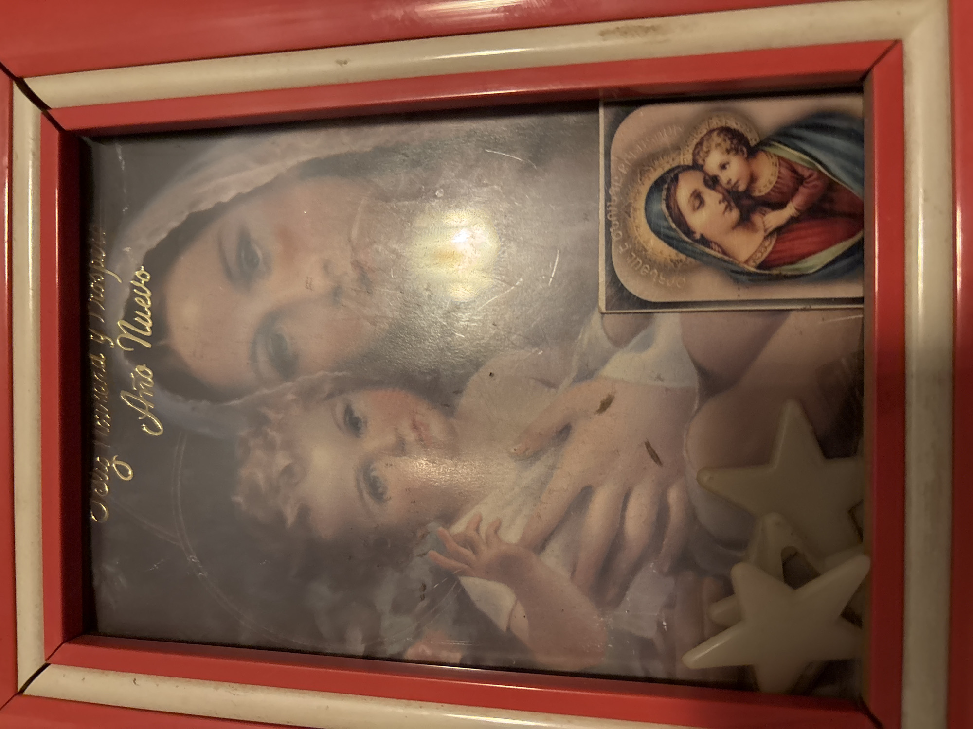 A framed image of the Virgin Mary and the baby Jesus hangs on a wall, probably in a house in Vagharshapat, Armenia. The image is protected by glass and shows wear and tear. A smaller, related image is attached to the top right corner of the main image. Two plastic stars adorn the bottom right corner of the frame. The frame is red with a white interior. The text "Feliz nuevo año" is on the left side of the picture.