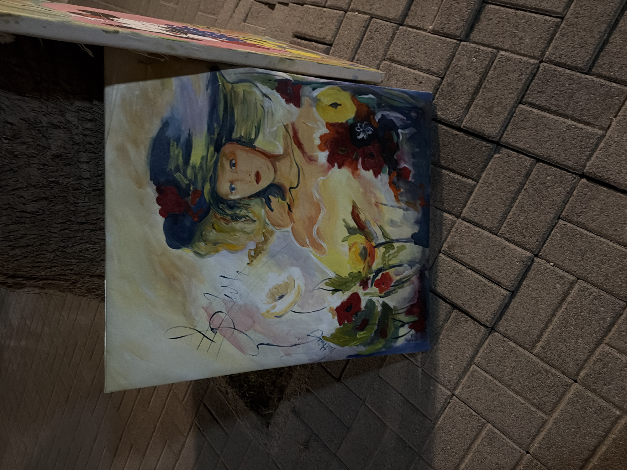 An oil painting of a woman wearing a wide-brimmed hat is displayed outdoors on a cobblestone sidewalk in Georgioupoli, Greece. The painting, which is propped up against a tree trunk, depicts the woman with blue eyes and red lips, surrounded by a colorful array of flowers. Her expression is serene. The painting is signed by the artist in the lower left corner. The surrounding environment is a pedestrian walkway with interlocking pavers.