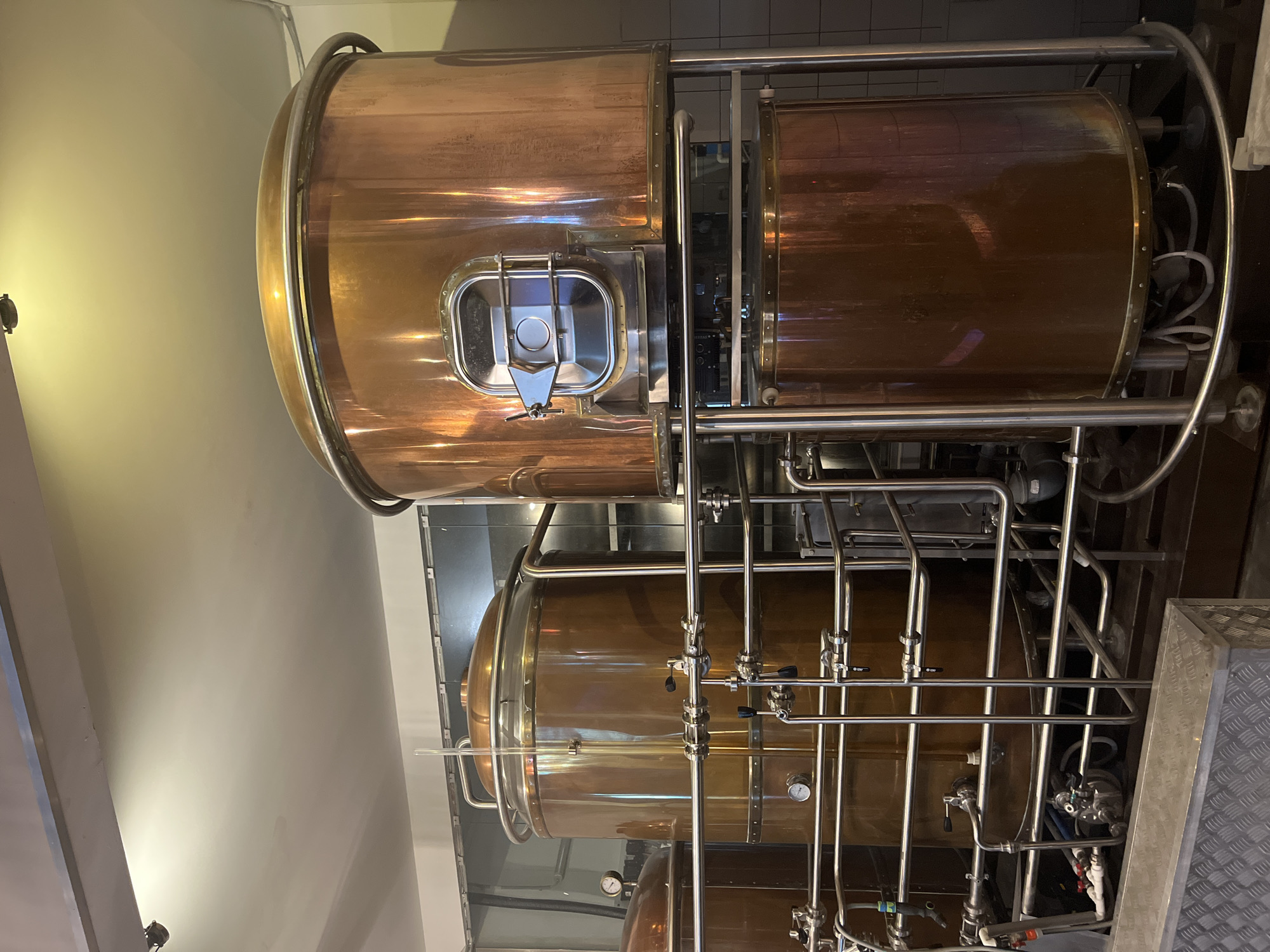The image shows a copper brewing system. There are two large copper tanks stacked vertically and supported by a stainless steel frame with pipes and valves. The tanks are located indoors, possibly in a brewery or distillery. The location could be Kazan, Russia, as requested. There are no people in the image. The lighting is bright and highlights the polished metal of the tanks and pipes.