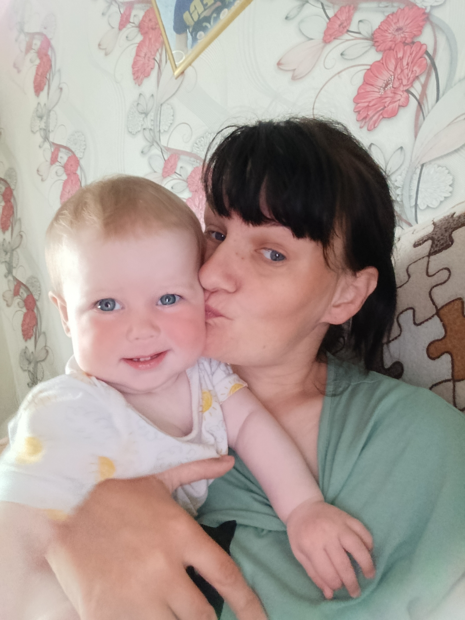 A photograph captures a woman kissing a baby on the cheek. The woman has dark hair and is wearing a green shirt. The baby has fair hair and blue eyes and is wearing a white shirt with yellow sun patterns. The background features wallpaper with a floral design and a framed picture. The context suggests an intimate moment between mother and child. The city is identified as Фоки, Russia, but the image lacks specific landmarks to confirm the exact location.