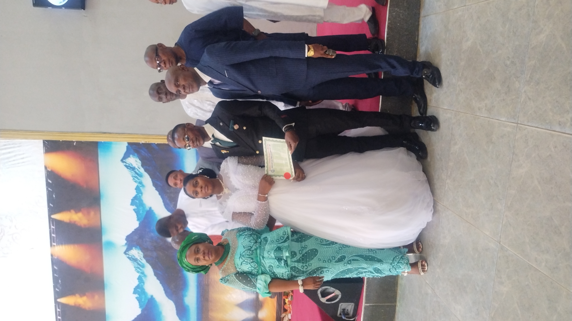 In a room with a concrete floor and a wall decorated with a backdrop featuring mountains and what appear to be flames, a wedding party poses for a photograph. In the foreground, a woman wearing a teal, beaded lace dress and a matching headscarf stands to the left of a bride in a white wedding gown. The bride holds a certificate in her hands. To the bride's right stands a groom in a dark suit, a green tie, and a white shirt. Behind them, several men in formal attire, including suits and traditional Nigerian clothing, are also visible. The overall scene suggests a formal wedding ceremony or reception taking place in Kabong, Nigeria.