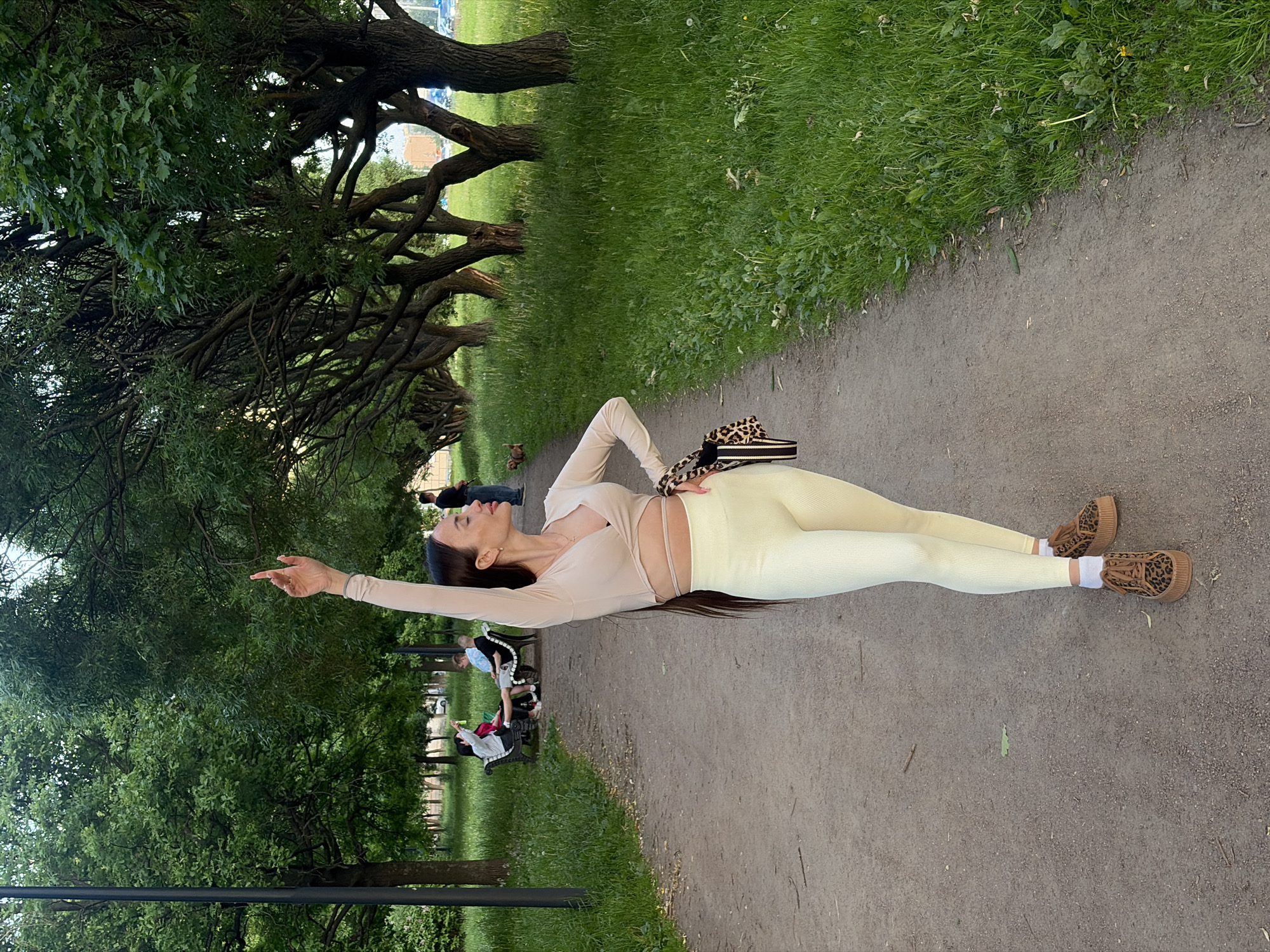 The image depicts an outdoor scene within a park or green area, likely situated within the city of Saint Petersburg, Russia, characterized by its mature trees and urban background elements. A wide, unpaved dirt path, light brown in color, runs through the center, receding into the distance.
The central subject is a slim woman standing on the path, positioned slightly to the right of the frame, facing right with her head tilted back and eyes closed. Her right arm is raised directly upward, with her hand slightly cupped and index finger pointing towards the sky, conveying a sense of looking or reaching. She wears a light beige, long-sleeved, ribbed crop top with a criss-cross front detail revealing a sliver of her midriff, paired with light yellow, ribbed, high-waisted leggings. On her feet are white socks and brown platform sneakers with a leopard print. Her long, dark brown hair cascades down her back. A black and leopard-print striped handbag hangs at her left hip.
To the left of the path, a dark lamppost stands near a cluster of dense green trees. Further back on the left, two individuals are seated on a white park bench beneath a canopy of deciduous trees. One person, wearing a light jacket, holds a red object, possibly a drink can, up in the air.
To the right of the path, a distinct row of mature trees with gnarled trunks and dense green foliage creates a natural barrier. Beyond these trees, a grassy area, partially overgrown with dandelions, extends. In the mid-ground on the right, a man in a blue shirt and dark pants stands facing away from the camera, and a small brown dog walks on the grass.
In the far background, urban structures, including multi-story buildings and a street with parked cars, are visible, indicating the park's proximity to an urban environment. The lighting is soft and even, suggesting either early morning or late afternoon.