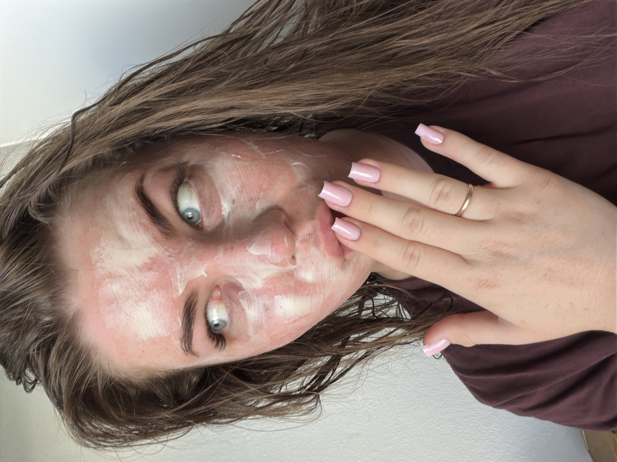 A woman with fair skin, light brown hair, and blue eyes is visible. She is wearing a brown t-shirt and has a white substance on her face, possibly a face mask. Her hand is near her face, displaying pink polished nails and a gold ring on her ring finger. The background appears to be a simple white wall. The image does not provide any information to determine its location in Chaikovsky, Russia.