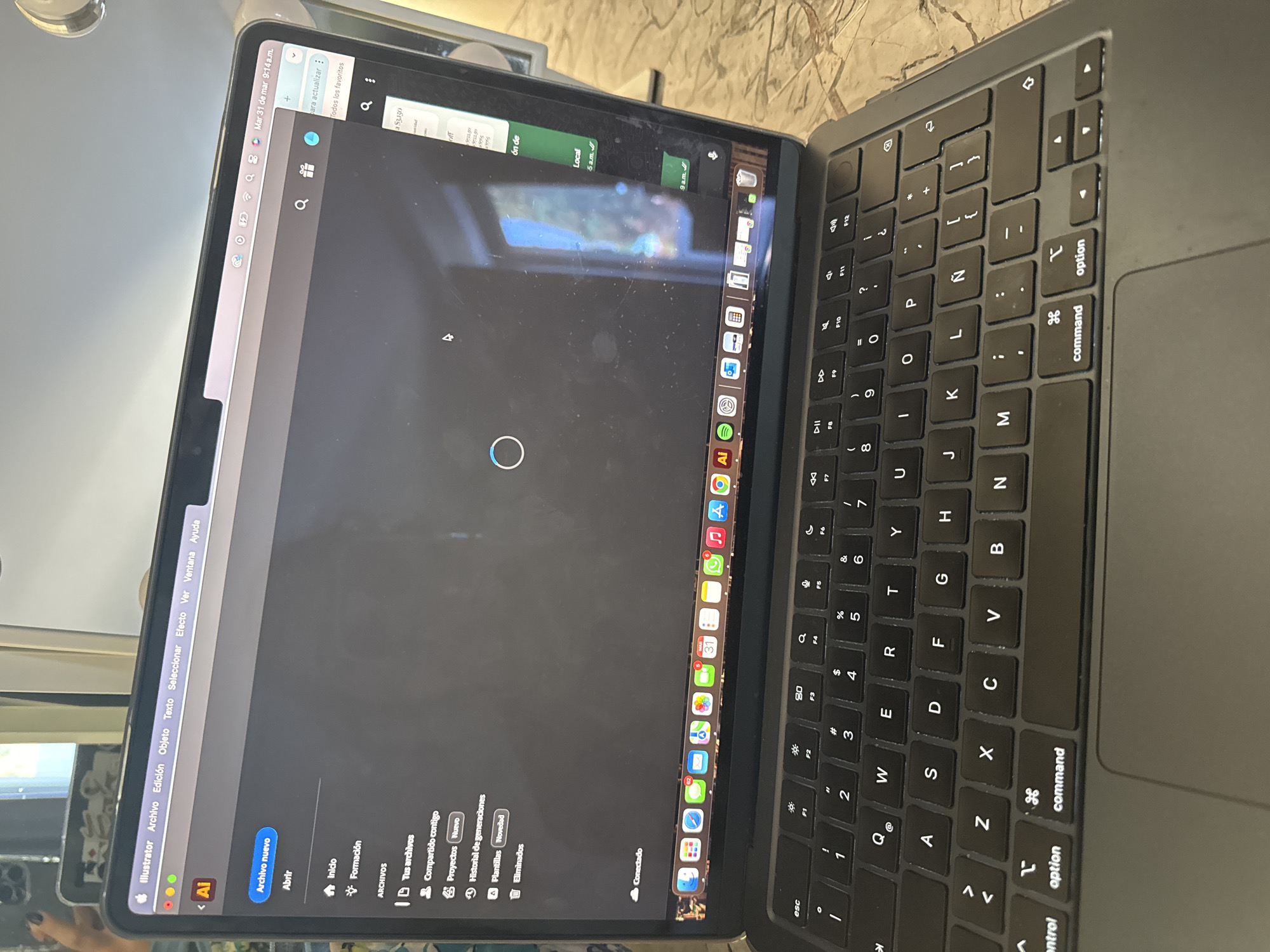 Here is a detailed description of the image:
**Scene:** Close-up view of a dark-colored Apple MacBook laptop, with the screen open and the keyboard visible. The laptop appears to be on a surface with a marble-like texture.
**Screen:** The screen displays the Adobe Illustrator application, likely in a Spanish-language version, as evidenced by the menu bar items such as "Archivo," "Edición," and "Objeto." A loading circle is in the center of the screen, indicating that the application is in the process of opening or accessing a file. The menu bar also shows the date "Mar 31 de mar 9:14a.m.". On the right side of the screen, there are several application icons displayed, as well as a pop up showing the word "Local 5 a.m."
**Keyboard:** The laptop keyboard is visible, showing the layout of keys and function keys.
**Environment:** The surface is mottled, and there are hints of other objects in the background, including what may be a window and some reflections, but the overall focus is on the laptop itself. There is a hand visible in the reflection of the screen.
**Location:** There is no visible information in the image to place the location in the city of San Bernardino Tlaxcalancingo, Mexico.