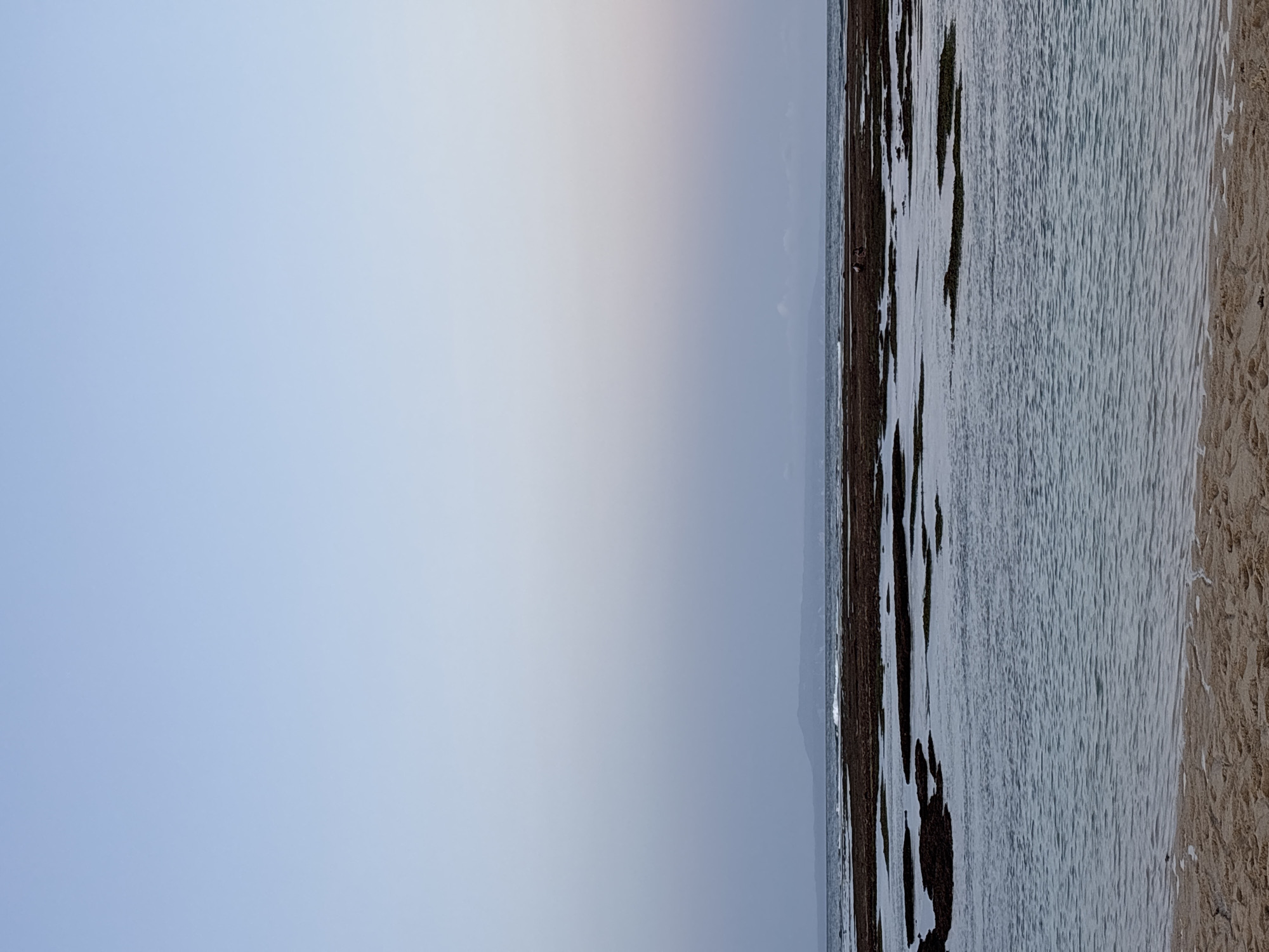 The image depicts a coastal scene, likely at low tide, in Kuta Selatan, Indonesia. The sky occupies the majority of the left portion of the frame, transitioning from a light blue near the top to a hazy gray toward the horizon. A thin strip of land is visible on the horizon line.
The right portion of the image shows the intertidal zone. The water appears shallow and rippled. Dark rocks, some covered with green algae, are interspersed within the water. To the right of the water is a sandy beach. The color of the sand is beige.
Two figures are visible on the shoreline. They are small relative to the overall scene and appear to be standing near the rocks. No other specific details about actions or interactions are evident.