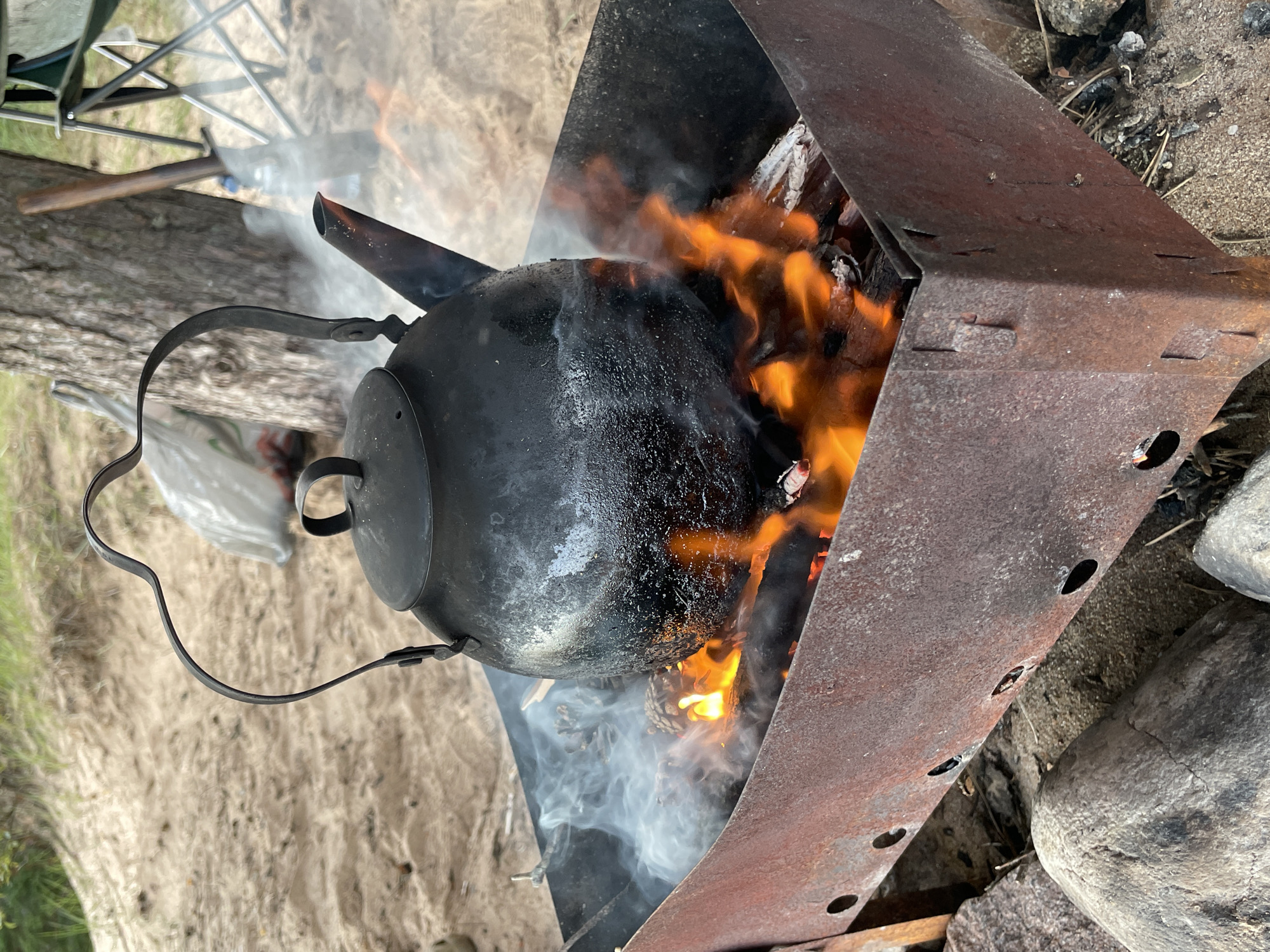 A close-up, eye-level shot captures a black metal kettle suspended over an open fire in a rustic, outdoor setting. The kettle, which appears to be made of cast iron, is filled with water and rests on a rusty metal brazier. Flames lick at the bottom of the kettle, producing smoke that billows upwards. The fire is fueled by wood and pinecones, adding to the primitive ambiance. Surrounding the brazier is a sandy surface, suggesting a beach or campsite. In the background, the natural landscape unfolds with sand dunes, vegetation, and a tree trunk. A folding chair and what appears to be a shovel lean against the tree, hinting at human presence and recreational activity. The scene is set in Zelenaya Roshcha, Russia, a location known for its natural beauty and outdoor recreational opportunities.