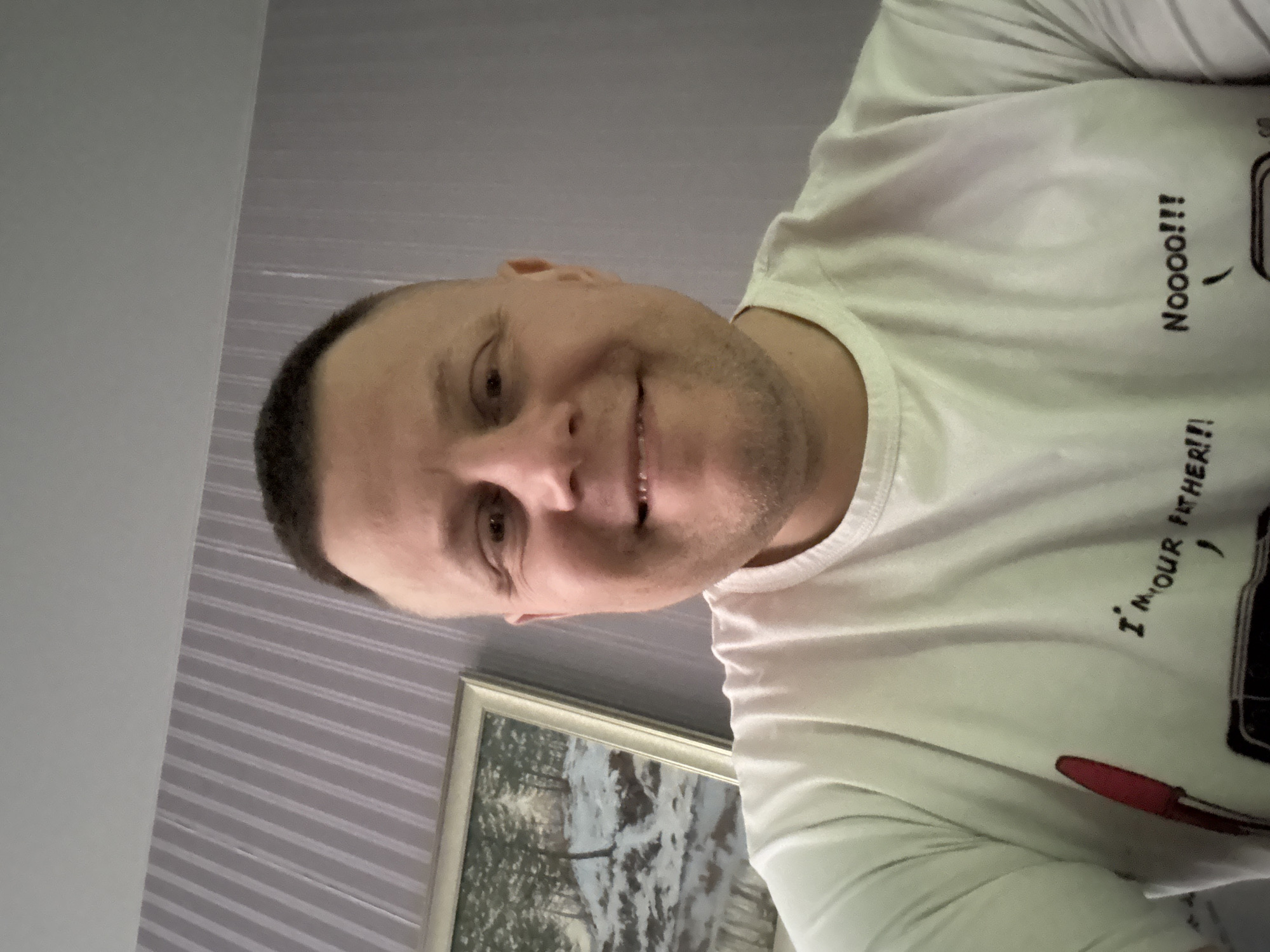 A selfie of a man with short, dark hair and fair skin. He is wearing a white t-shirt with black writing and a drawing of a cassette player with "I'm Your Father!" and "Noooo!!!" written next to it. He is smiling. Behind him is a wall with striped wallpaper and a framed picture. The location is indicated as Minsk, Belarus, though there are no clear features in the image to confirm the exact location.