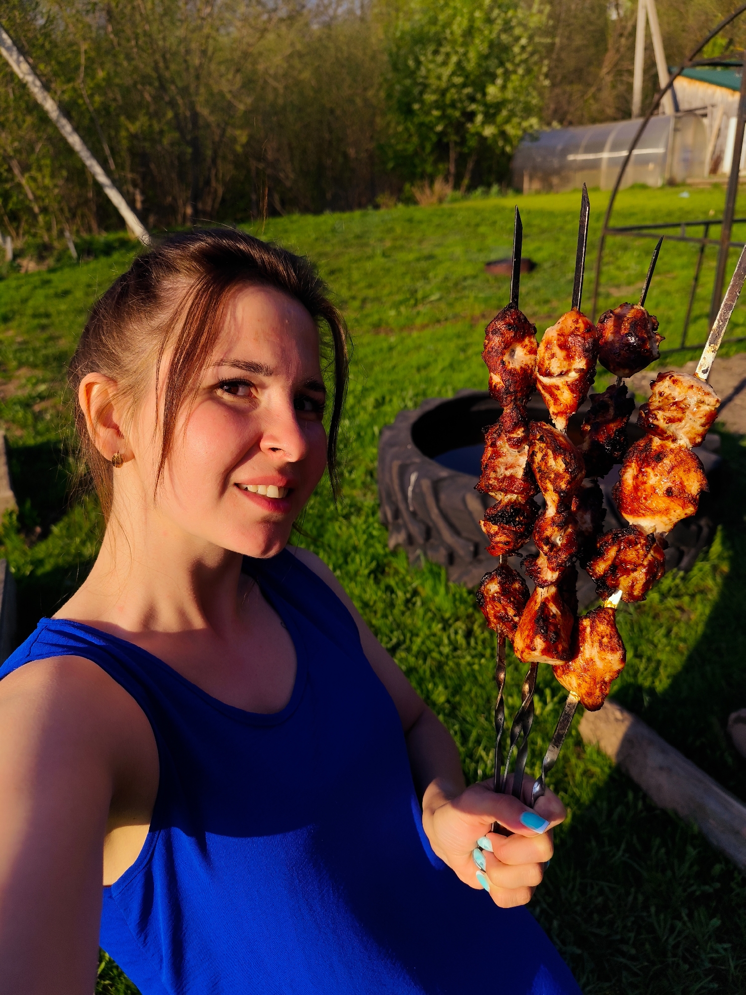 A woman is smiling at the camera while holding three skewers of cooked meat in her right hand. She has brown hair pulled back in a ponytail, fair skin, and is wearing a blue tank top. Her fingernails are painted blue. Behind her, there is a green lawn, trees, and a greenhouse. A large black tire sits on the ground behind the skewers. The photo appears to have been taken outdoors on a sunny day in Фоки, Russia.