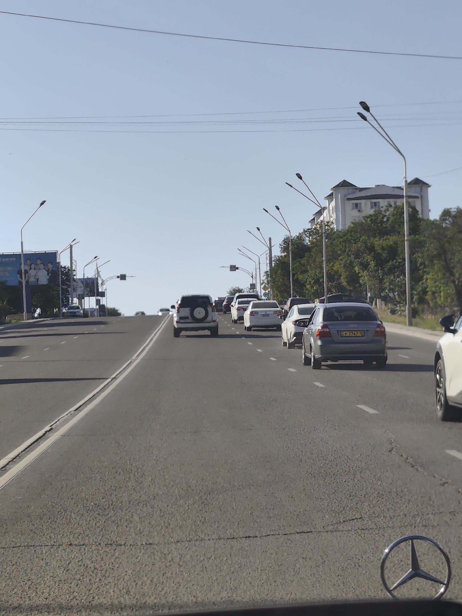 A forward-facing, eye-level shot shows a multi-lane road with traffic. The sky is clear and blue, indicating it's daytime, likely late morning or early afternoon due to the bright sunlight casting shadows. Several vehicles are visible, including SUVs and sedans, driving in the same direction. The road is paved with asphalt and marked with white lines. On the left side of the road, there are billboards and streetlights. On the right, there are trees and a large, modern building with a distinctive architectural style. The overall scene appears to be a suburban or urban setting. A Mercedes-Benz emblem is visible in the foreground, suggesting the photo was taken from inside a Mercedes-Benz vehicle. No specific activity or event is clearly depicted beyond general traffic flow. There is no visible text that can be read clearly.
