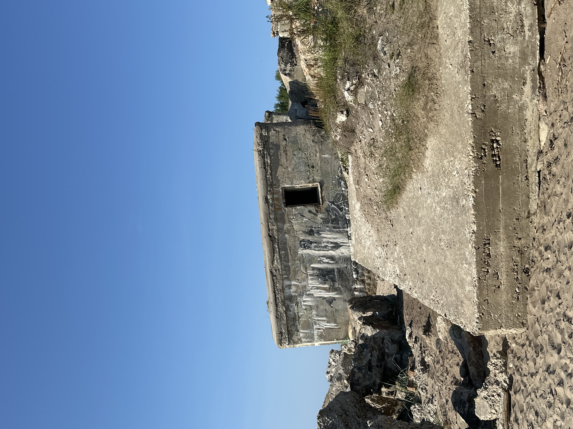 The image depicts a dilapidated concrete structure, possibly a bunker or an old military fortification, situated on a sandy, rocky terrain. The structure has a weathered, graffiti-covered facade with an open, dark window. Surrounding it are piles of rubble and sparse vegetation. In the foreground, a large, rectangular concrete slab rests on the sand. The scene is under a clear, bright blue sky, suggesting daytime. This location is the Liepaja Northern Forts, also known as the Karosta fortifications, in Liepaja, Latvia.
