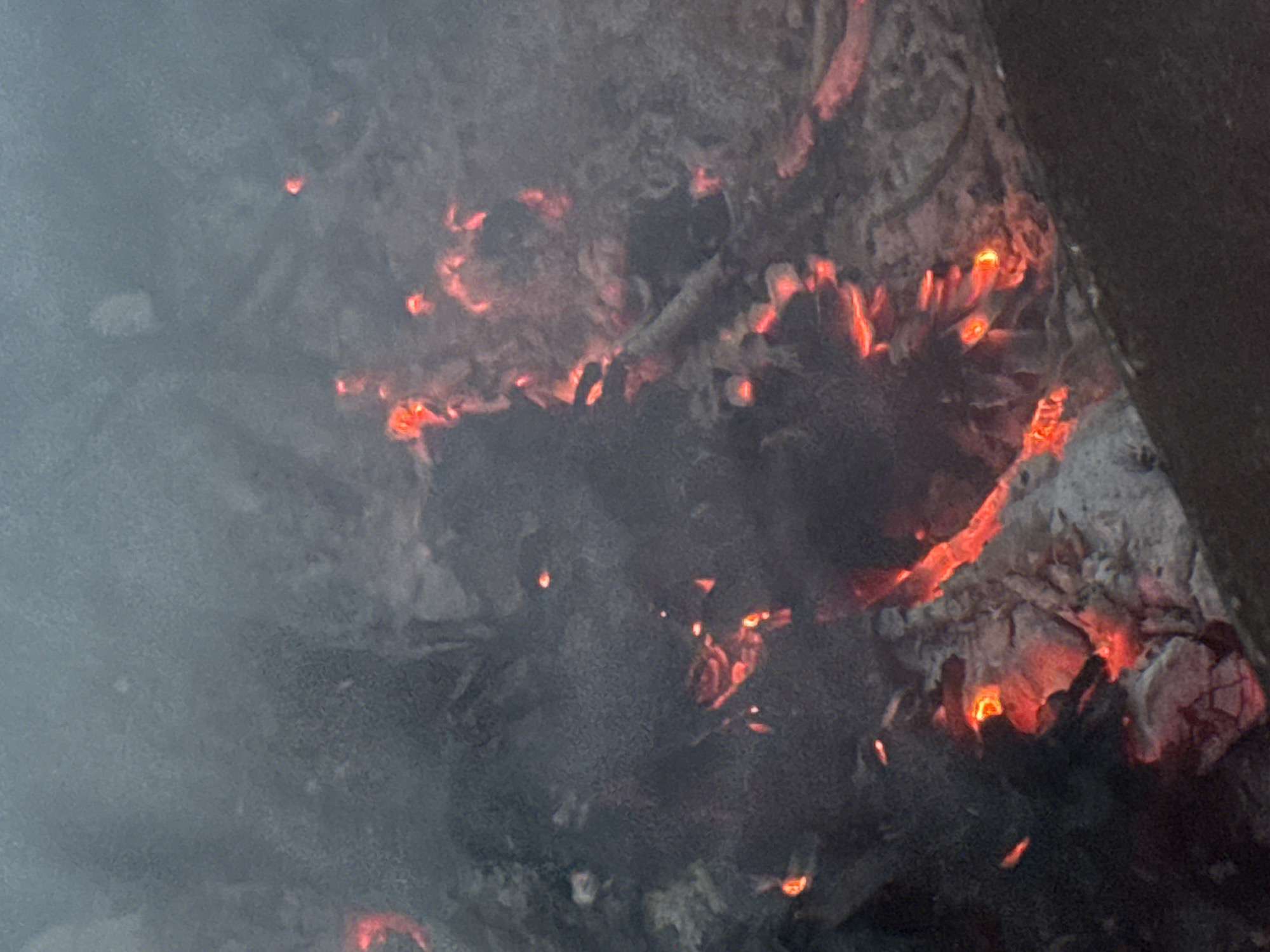 The image shows a close-up view of a dying fire in a dark, smoky environment. Embers glow with a vibrant orange-red hue against a backdrop of ash and charred material. Wisps of smoke drift upwards, obscuring the details of the scene. The texture of the ash is rough and uneven, with some larger pieces of wood or debris visible. The overall impression is one of warmth and fading light, with a sense of residual heat from the fire. The location in the city of Shepelevo, Russia, is not discernible from the image itself.
