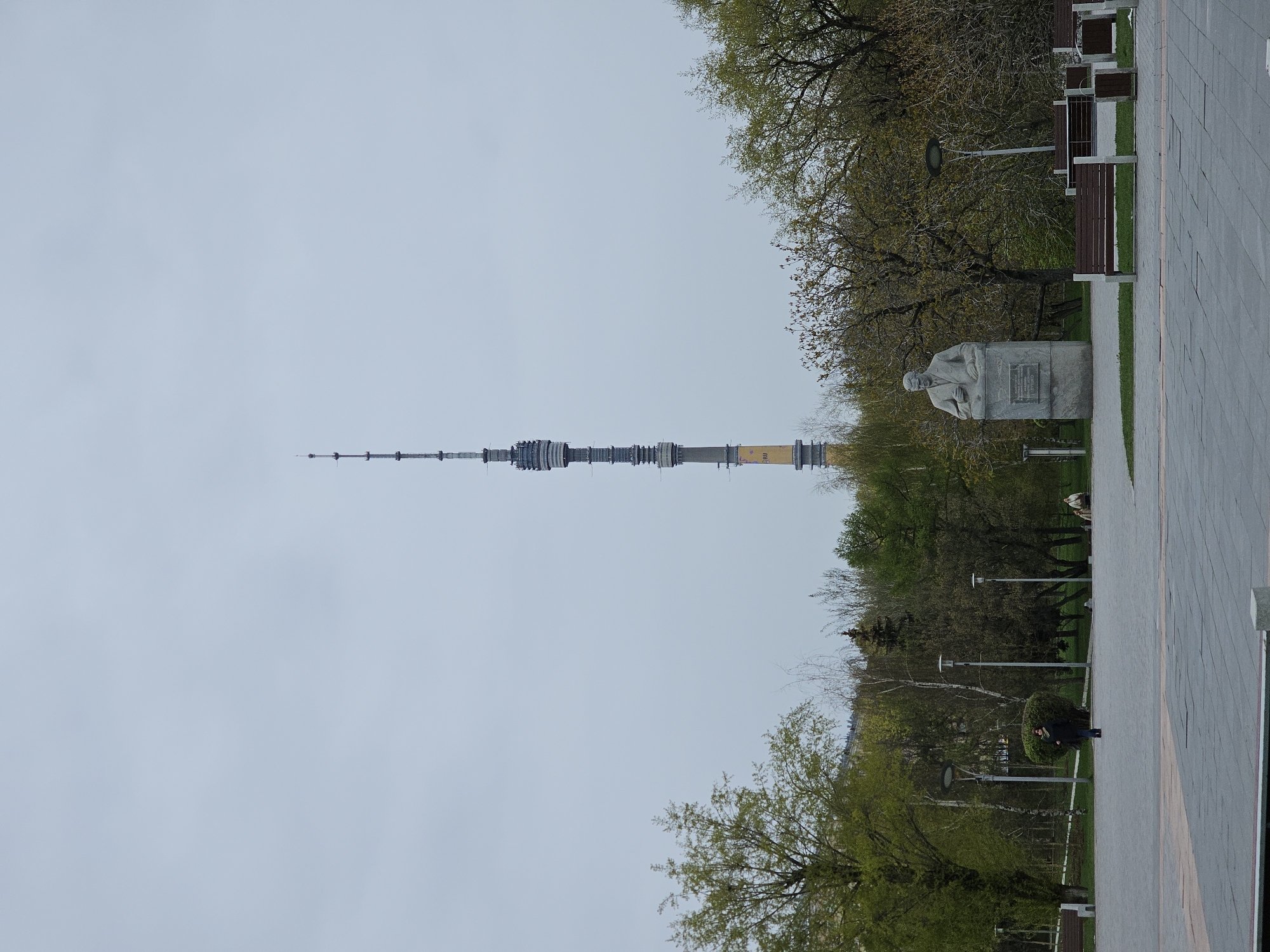 The image captures a panoramic view of a public park in Moscow, Russia, with the iconic Ostankino Television Tower prominently featured on the left. The sky is a soft, uniform light gray, indicating an overcast or cloudy day.
The Ostankino Tower dominates the left half of the frame, a slender and tall structure extending vertically towards the sky. Its shaft is characterized by a segmented, cylindrical design, with varying shades of gray, white, and a distinct yellow-orange section visible mid-way up its visible portion. A fine antenna spire tops the structure.
On the right side of the image, a paved walkway, composed of light-colored square paving stones, runs parallel to the tower. Adjacent to the walkway is a green lawn interspersed with trees. Some trees exhibit full, vibrant green foliage, indicative of spring or early summer, while others have sparser branches, possibly showing new growth. Along the pathway, several dark-brown park benches with metallic frames are positioned facing the park. Further into the park, a stone statue of a man is situated on a pedestal. The figure appears to be a bust or full-body statue of a man in formal attire, though details are indistinct. A rectangular plaque is visible on the pedestal, but any text on it is illegible due to the distance and resolution. Streetlights or lampposts with light-colored poles are also visible within the park area.
The overall scene depicts a serene urban park environment, devoid of specific human activity, under a subdued light.