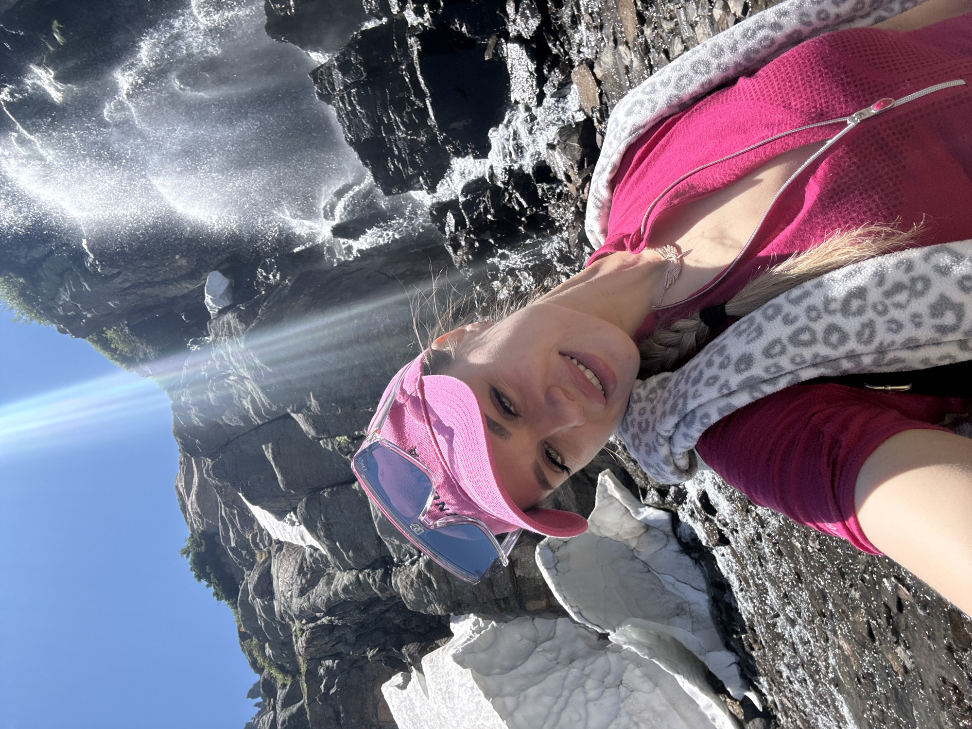 In a selfie taken outdoors, a woman with blonde hair braided down her side smiles at the camera. She is wearing a bright pink baseball cap and oversized sunglasses. A pink zip-up shirt is visible, along with a gray and white leopard-print jacket draped over her shoulders. She has a delicate necklace with a winged pendant. Behind her, a waterfall cascades down a rocky cliff face. Patches of snow are visible on the rocks and ground. The sun shines brightly, casting lens flares across the image. The scene appears to be in a mountainous area, possibly near Allenspark, Colorado, given the resemblance to the natural landscape.
