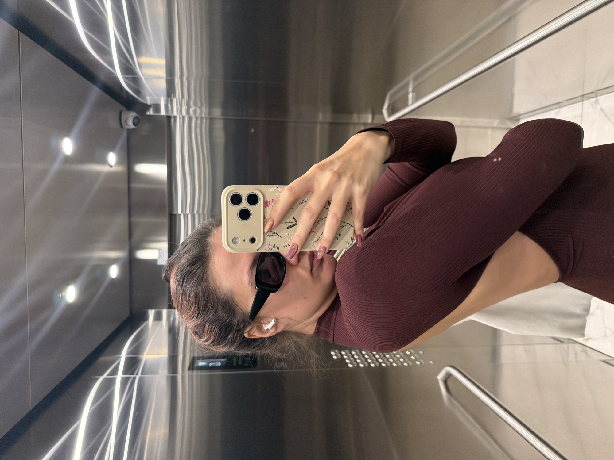 A woman stands in an elevator, taking a selfie with a phone that has a floral case. She wears a long-sleeved maroon shirt and sunglasses. The elevator is stainless steel, with a surveillance camera in the upper left corner. The location is likely Belgrade, Serbia, as indicated by the surrounding environment.