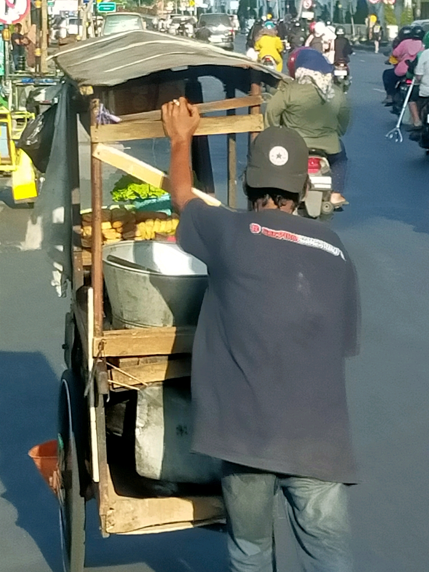 In Surabaya, Indonesia, a street vendor pushes a food cart on a paved road. The vendor, wearing a dark blue t-shirt with a red and white logo and blue jeans, has their right hand on a wooden bar of the cart. The cart has a dark grey tarp covering its top and is laden with various items, including a large metal basin, what appear to be fried foods, and some green leafy vegetables. The cart is partially obscured by the vendor's body and some black plastic sheeting. In the background, several other people are seen on motorcycles, some wearing colorful clothing and helmets, indicating a busy urban environment. The road surface is visible, and there are shadows cast by the objects in the scene.